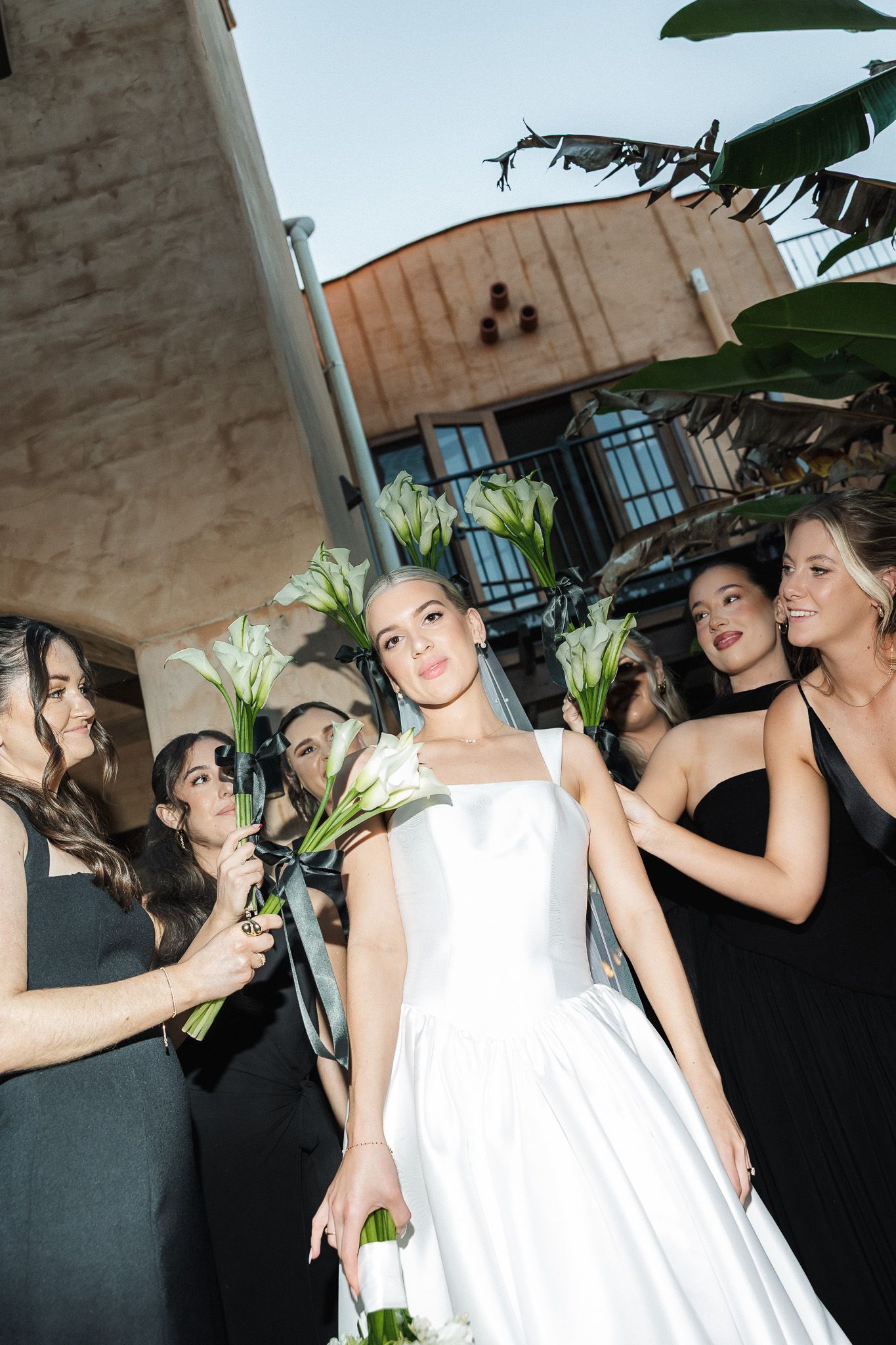 Bride in white gown holds calla lily bouquet surrounded by bridesmaids in black dresses at outdoor wedding venue.