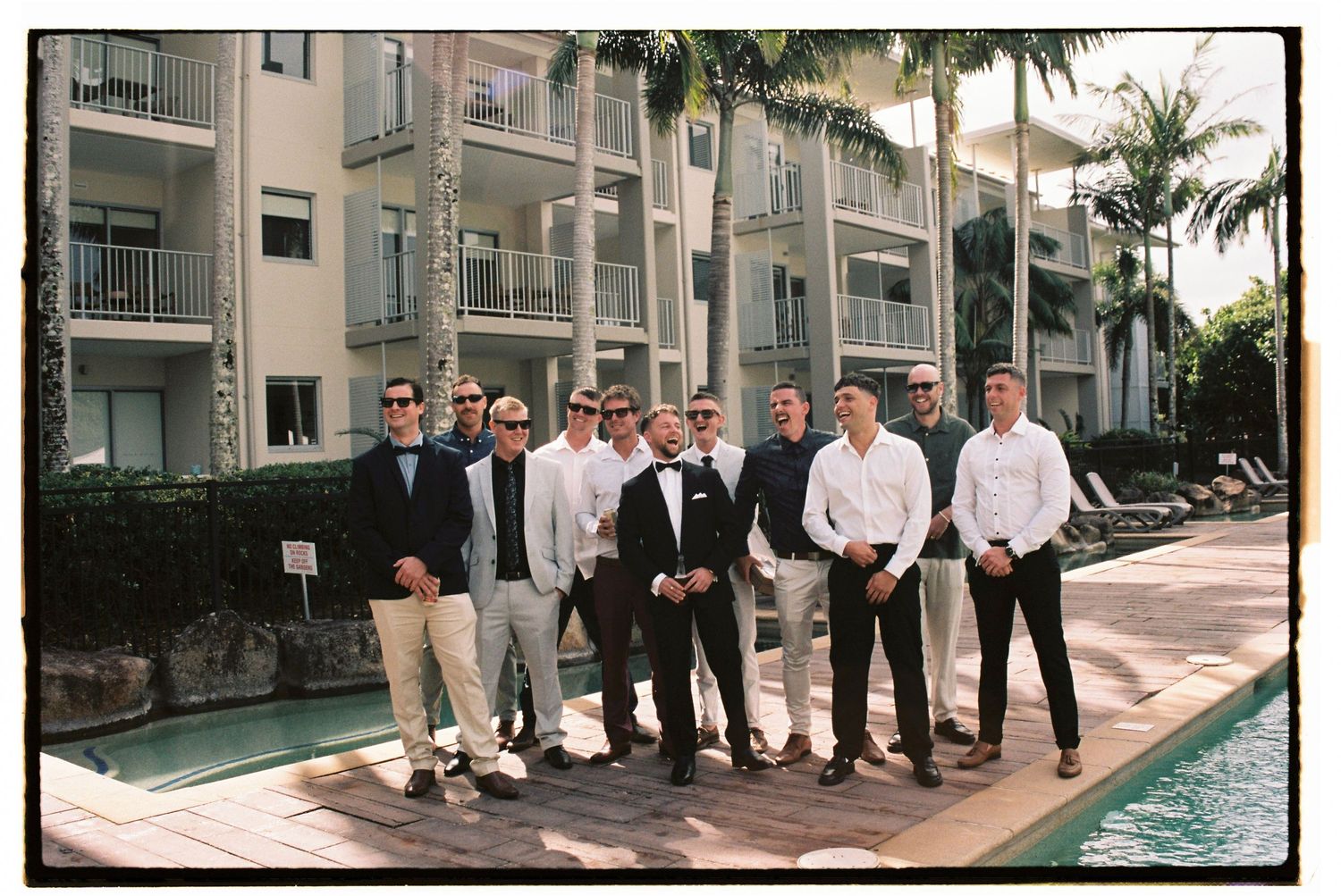 Groom and groomsmen pose poolside at tropical resort venue before wedding.
