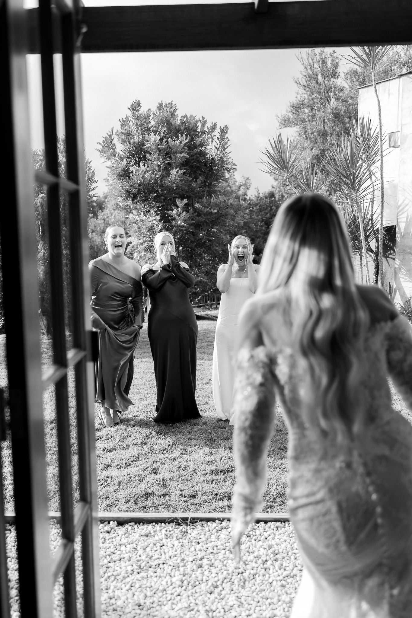 Black and white photo of a bride revealing her wedding dress to emotional bridesmaids outdoors.
