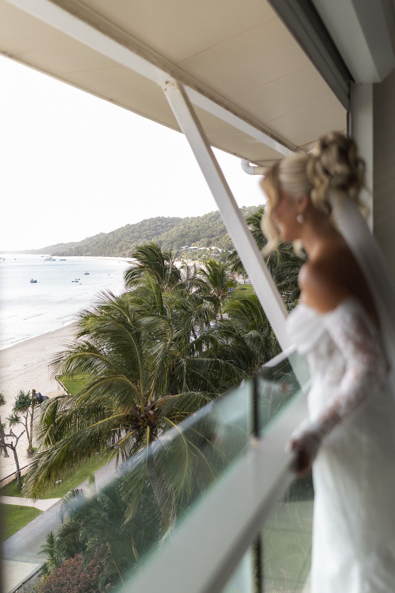 Bride in white lace gown gazes from balcony overlooking tropical beach with palm trees and ocean views.