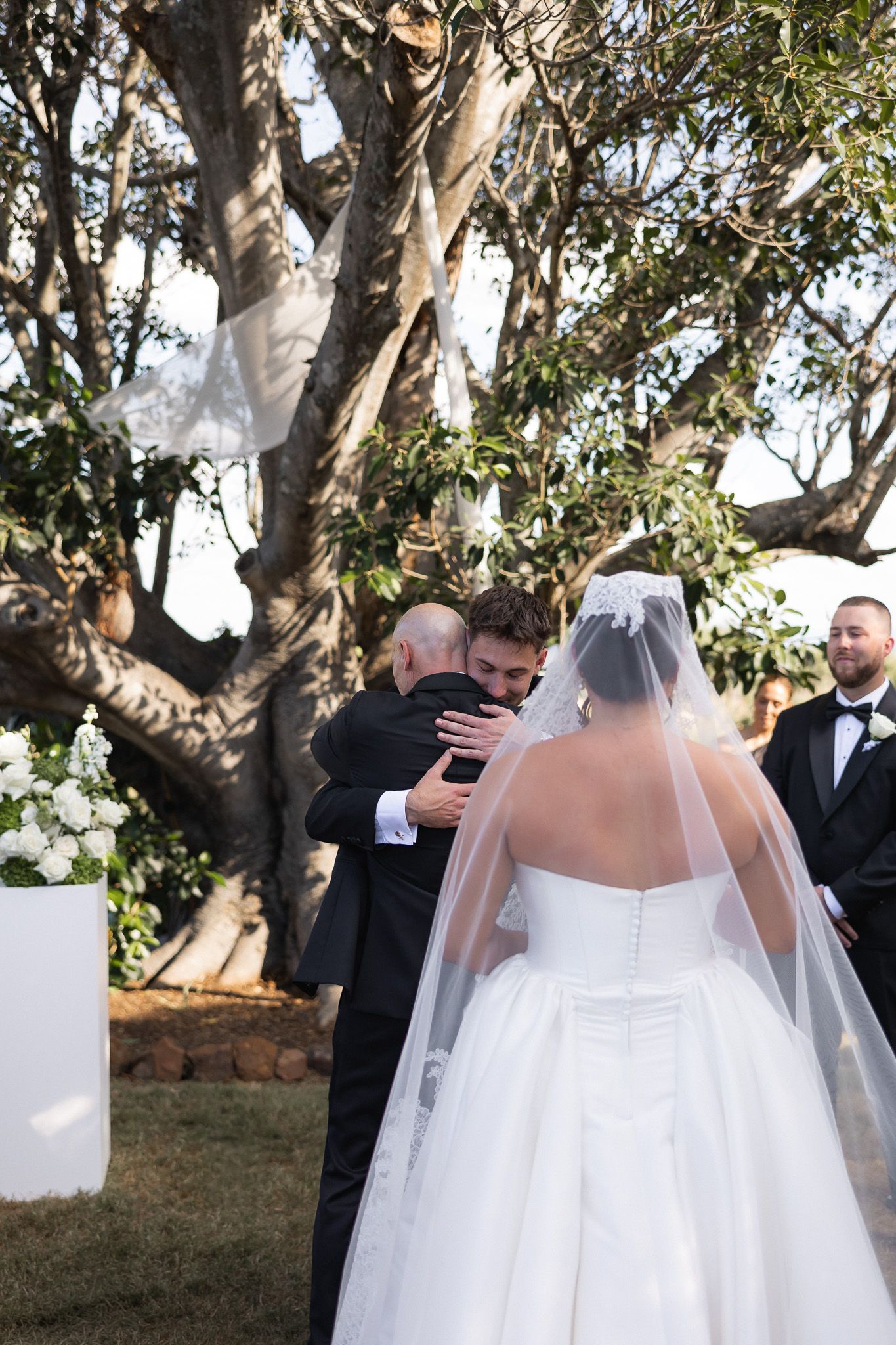 Bride receives emotional hug from father at outdoor wedding ceremony beneath large tree.