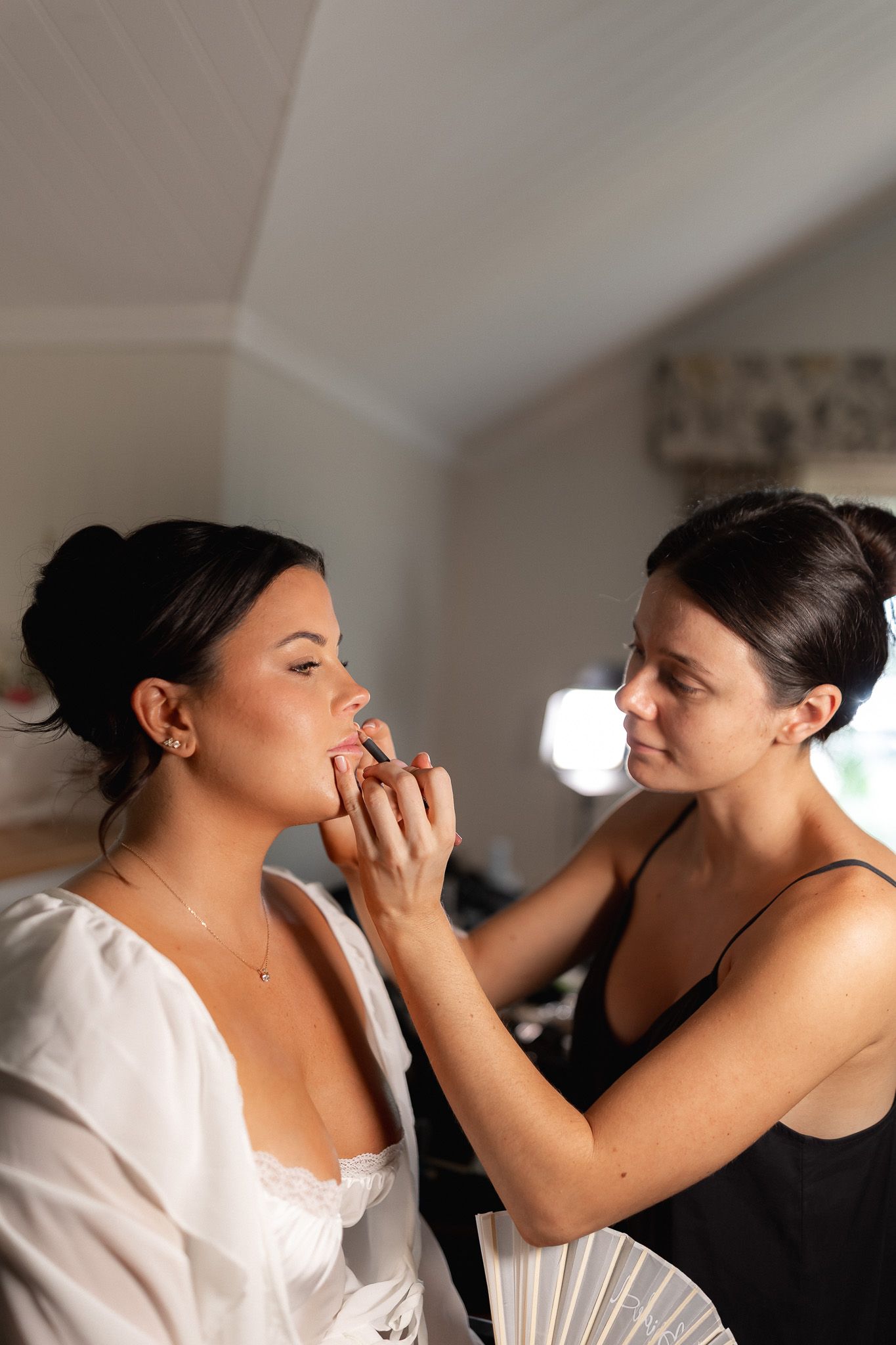 A makeup artist applying lipstick to a bride in a white robe during wedding preparations indoors.