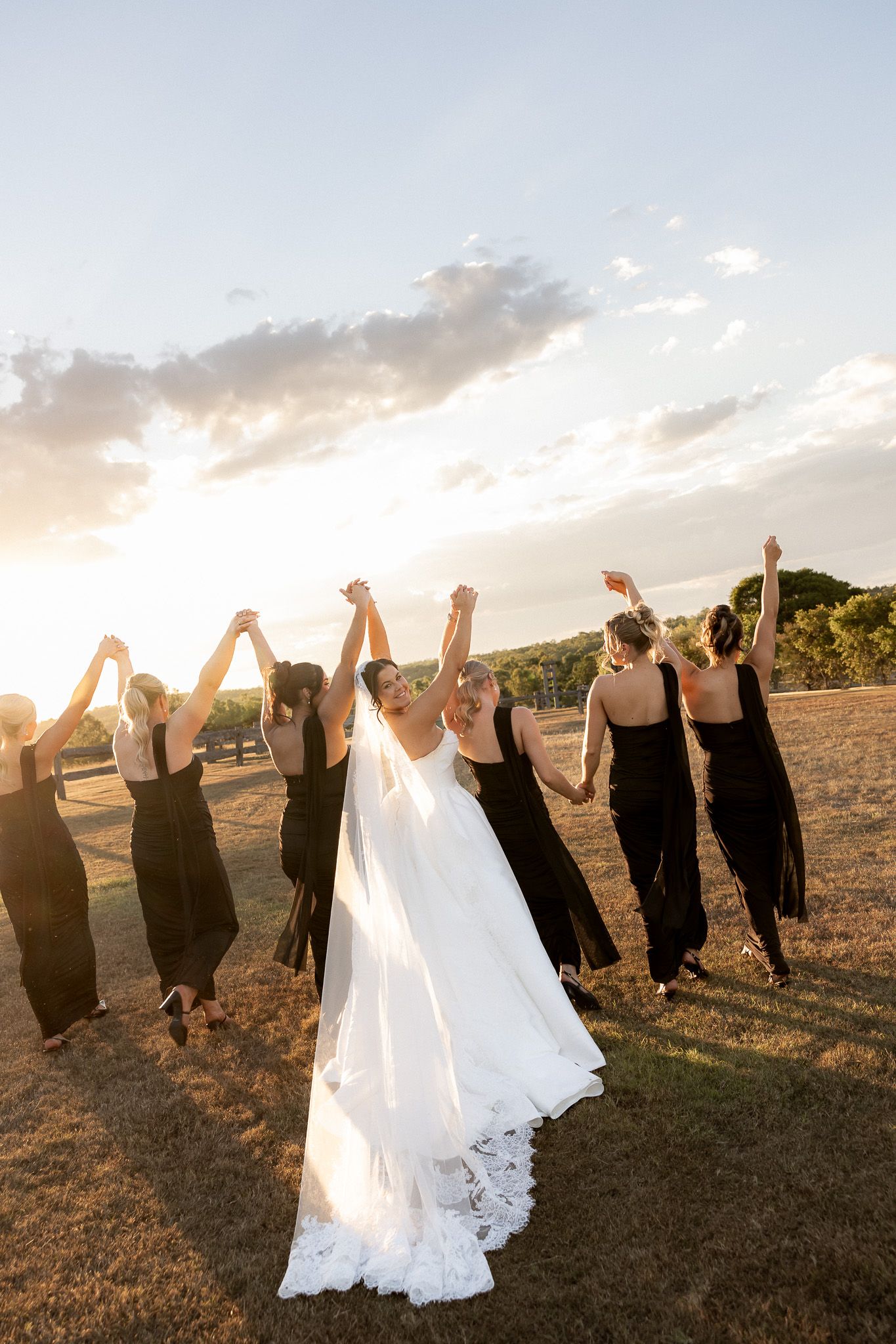 Bride in white lace gown holds hands with bridesmaids in black dresses at golden sunset in open field.
