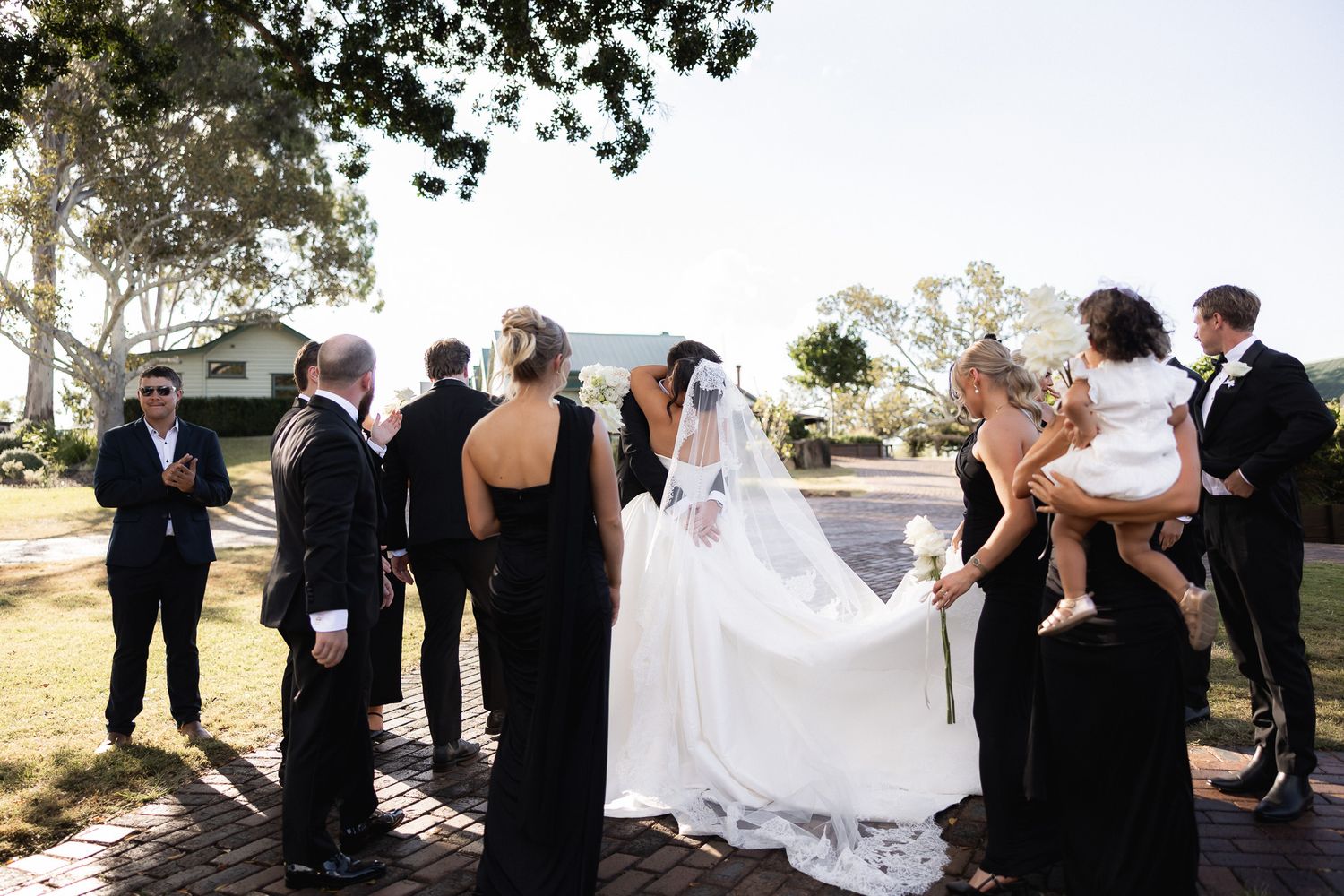 Wedding party in black and white attire gathered on brick path with bride in white gown.