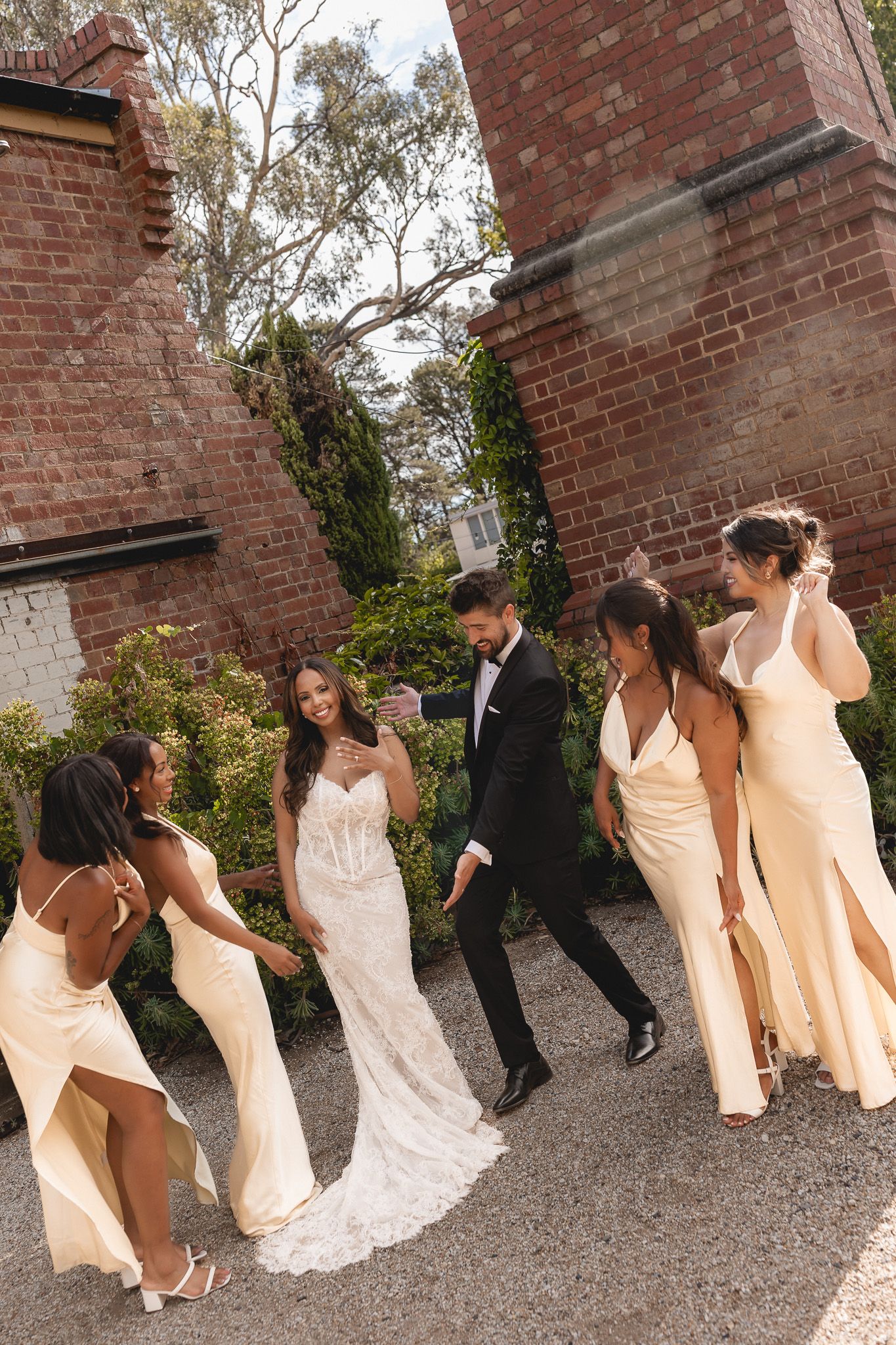 Bride and groom walking with bridesmaids in champagne gowns beside rustic brick walls and greenery.