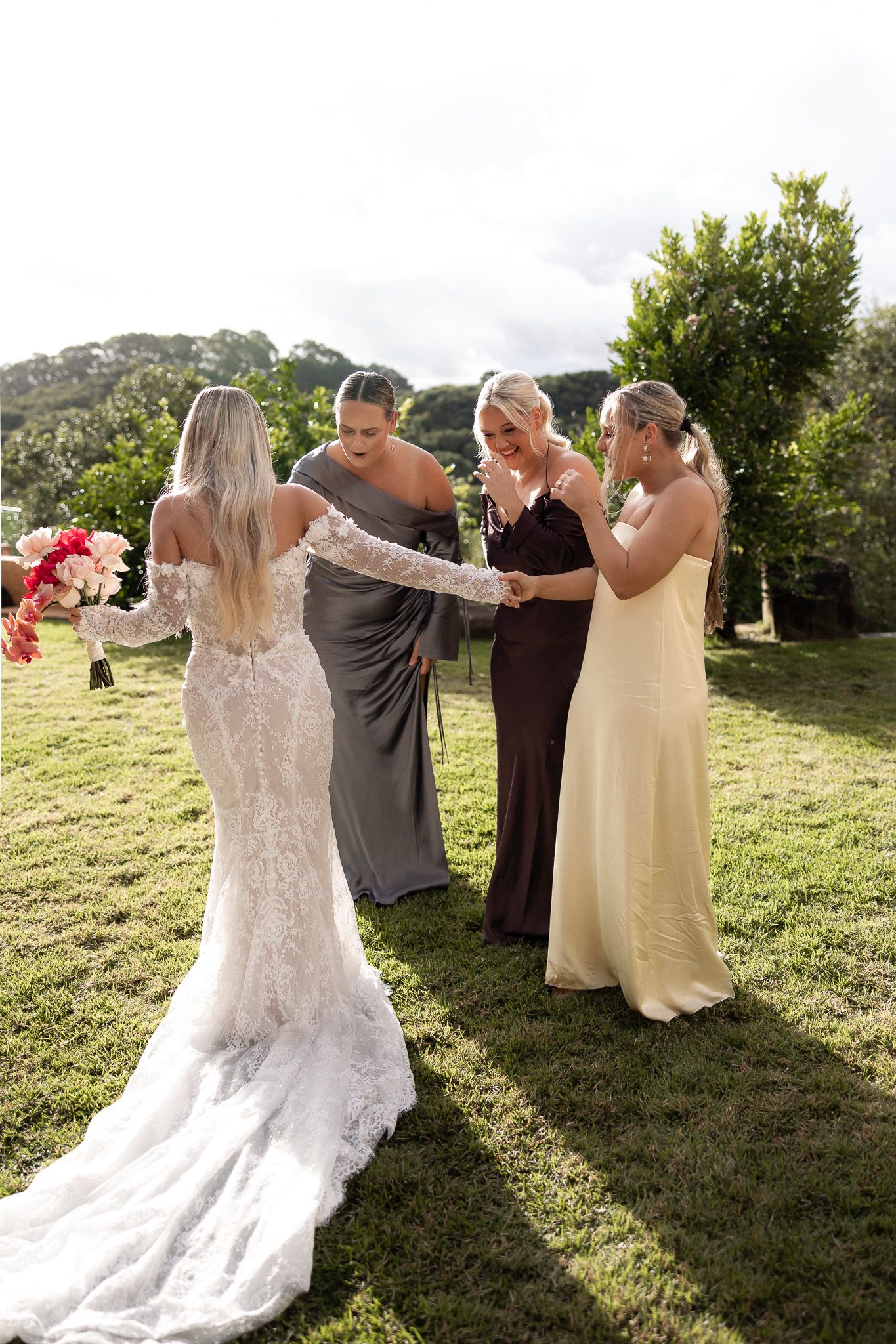 Bride in lace gown with bridesmaids in gray, brown, and yellow dresses gathered on green lawn.
