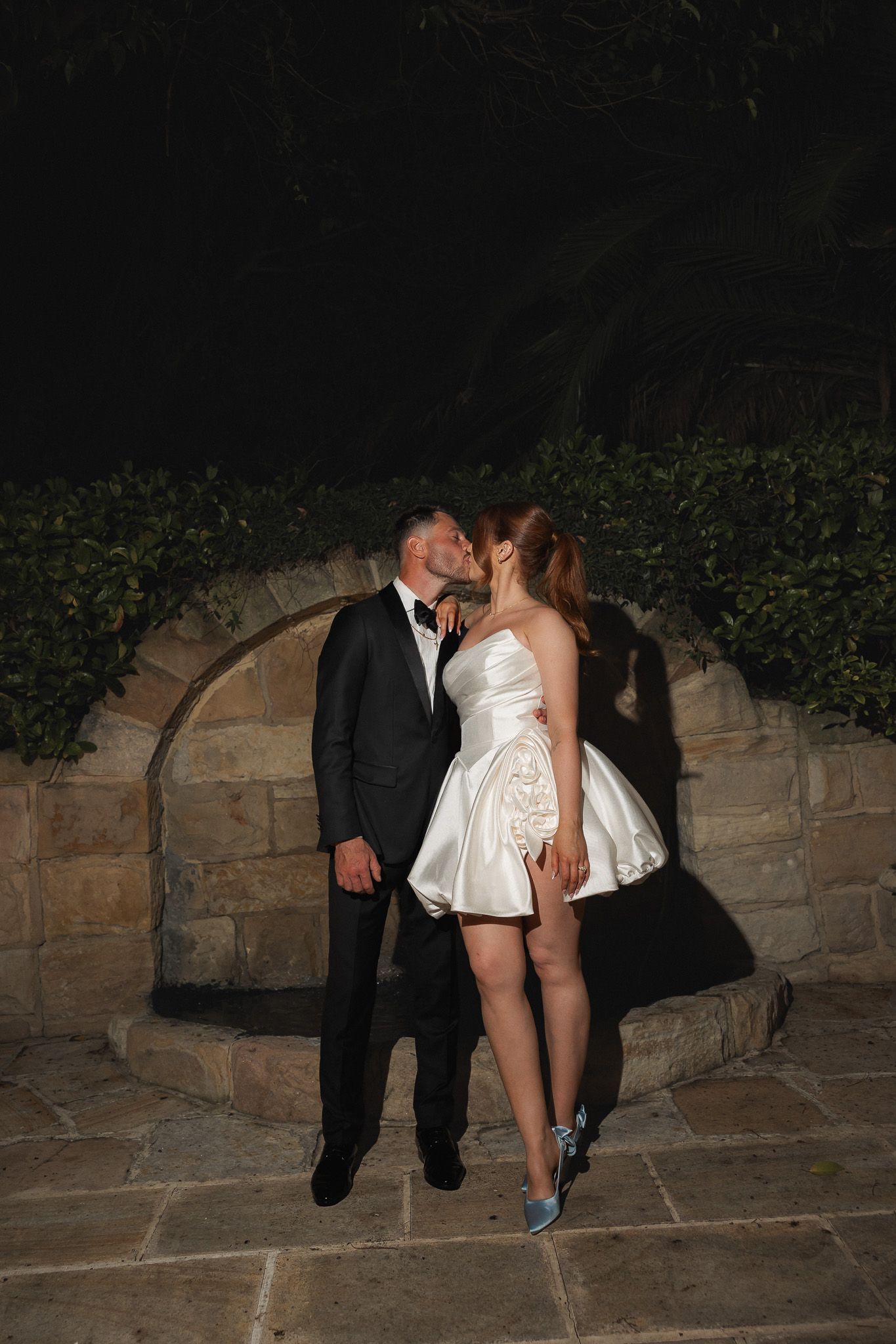 Couple sharing a romantic kiss at night, man in tuxedo and woman in white mini dress, standing by stone arch wall.