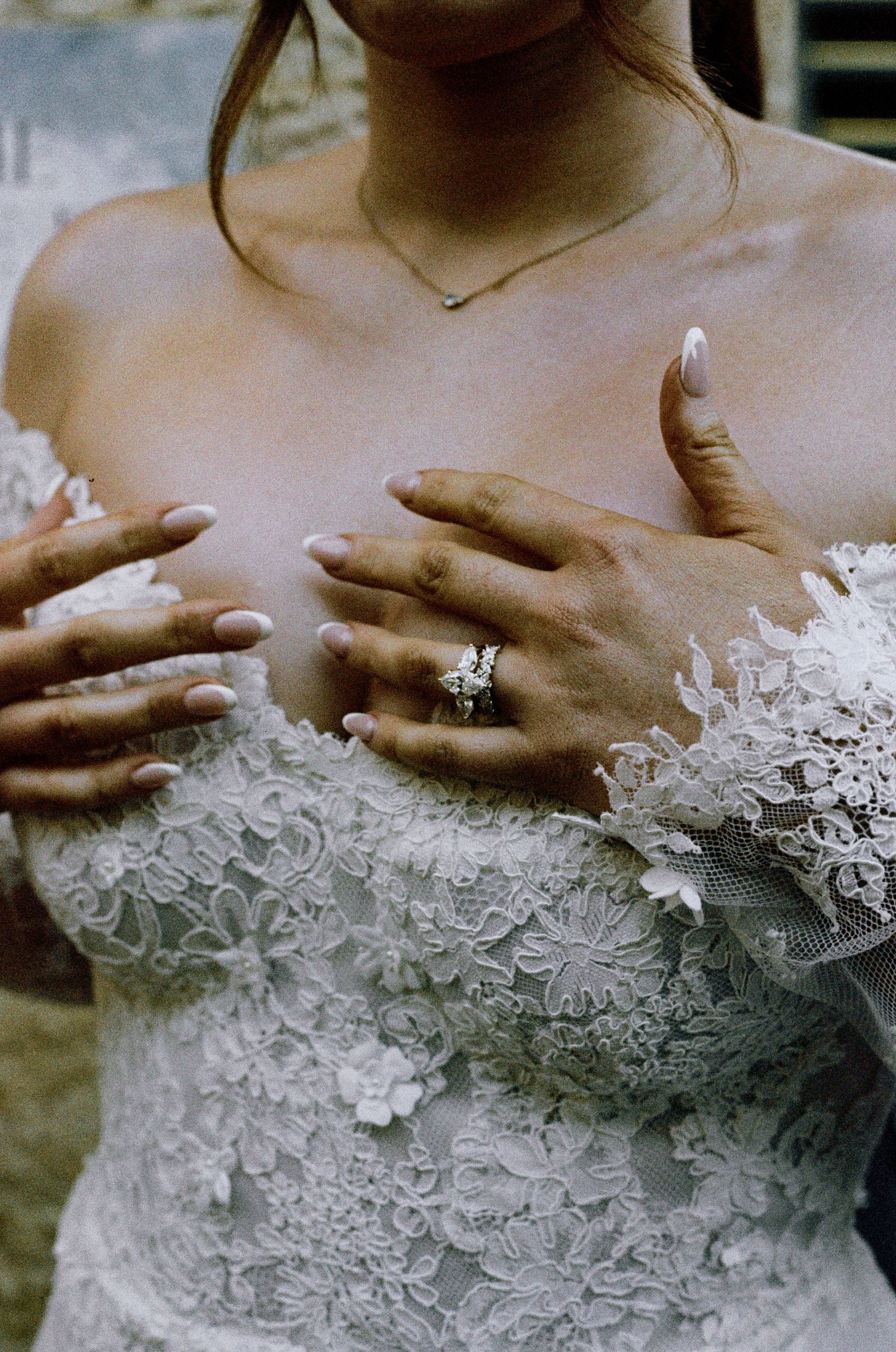 Bride in lace strapless wedding gown adjusting dress, showcasing diamond engagement ring and delicate necklace.