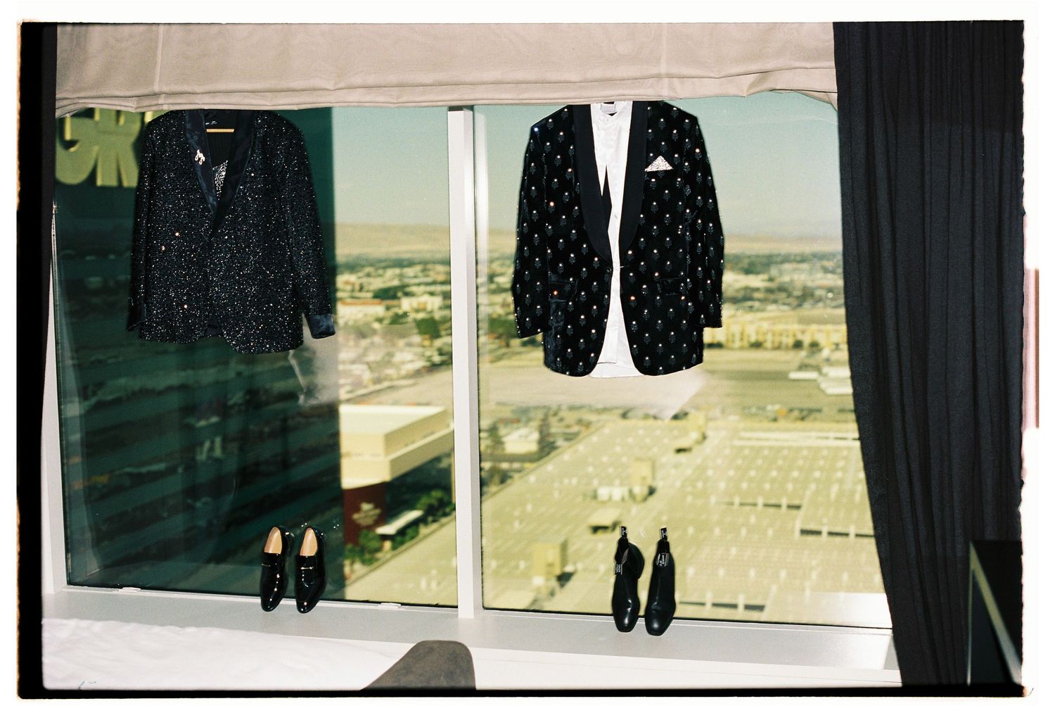 Two patterned suits and dress shoes displayed hanging in a window with a city skyline view below.