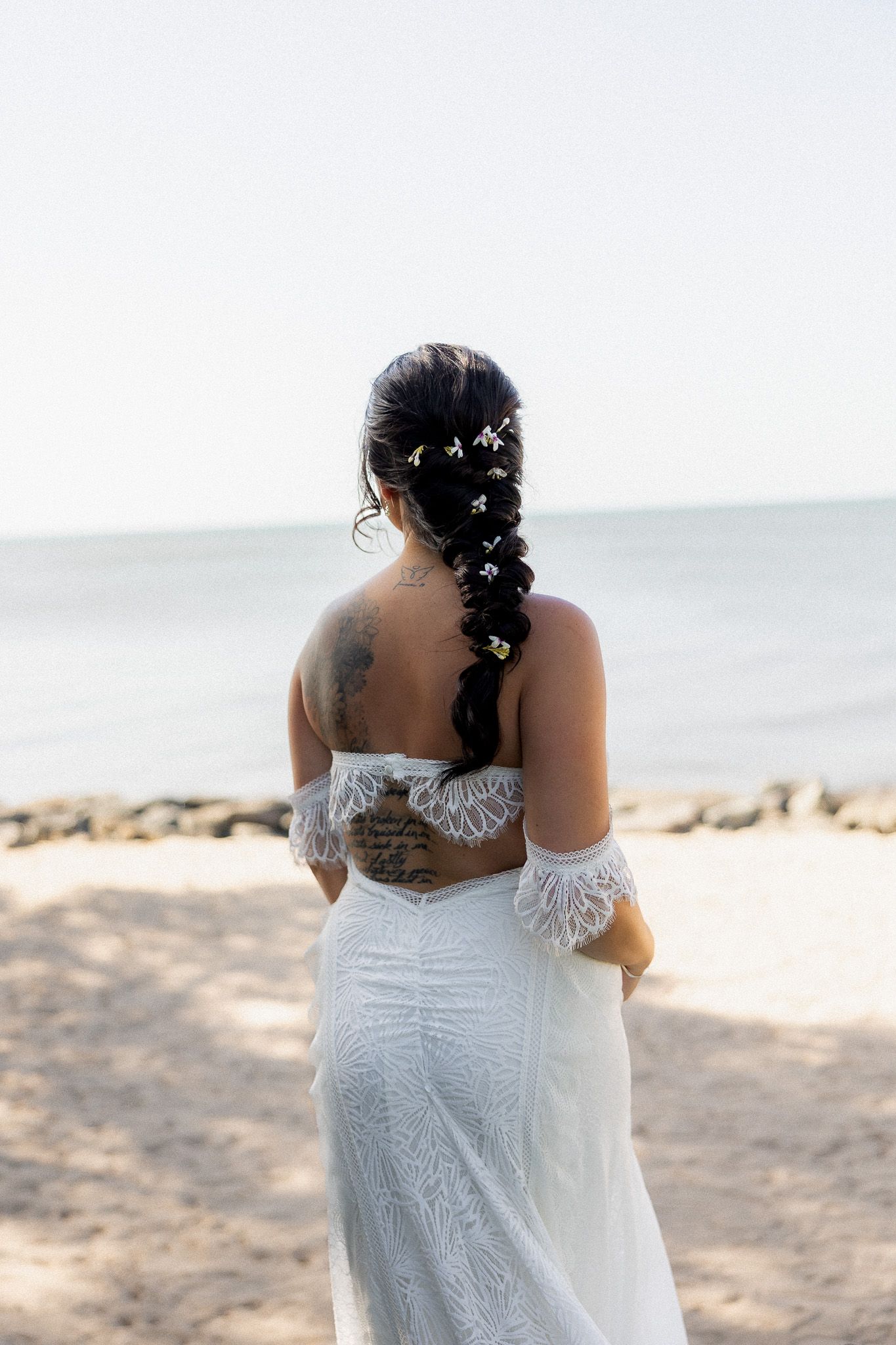 Bride with braided hair adorned with flowers and back tattoo stands on beach in lace gown.