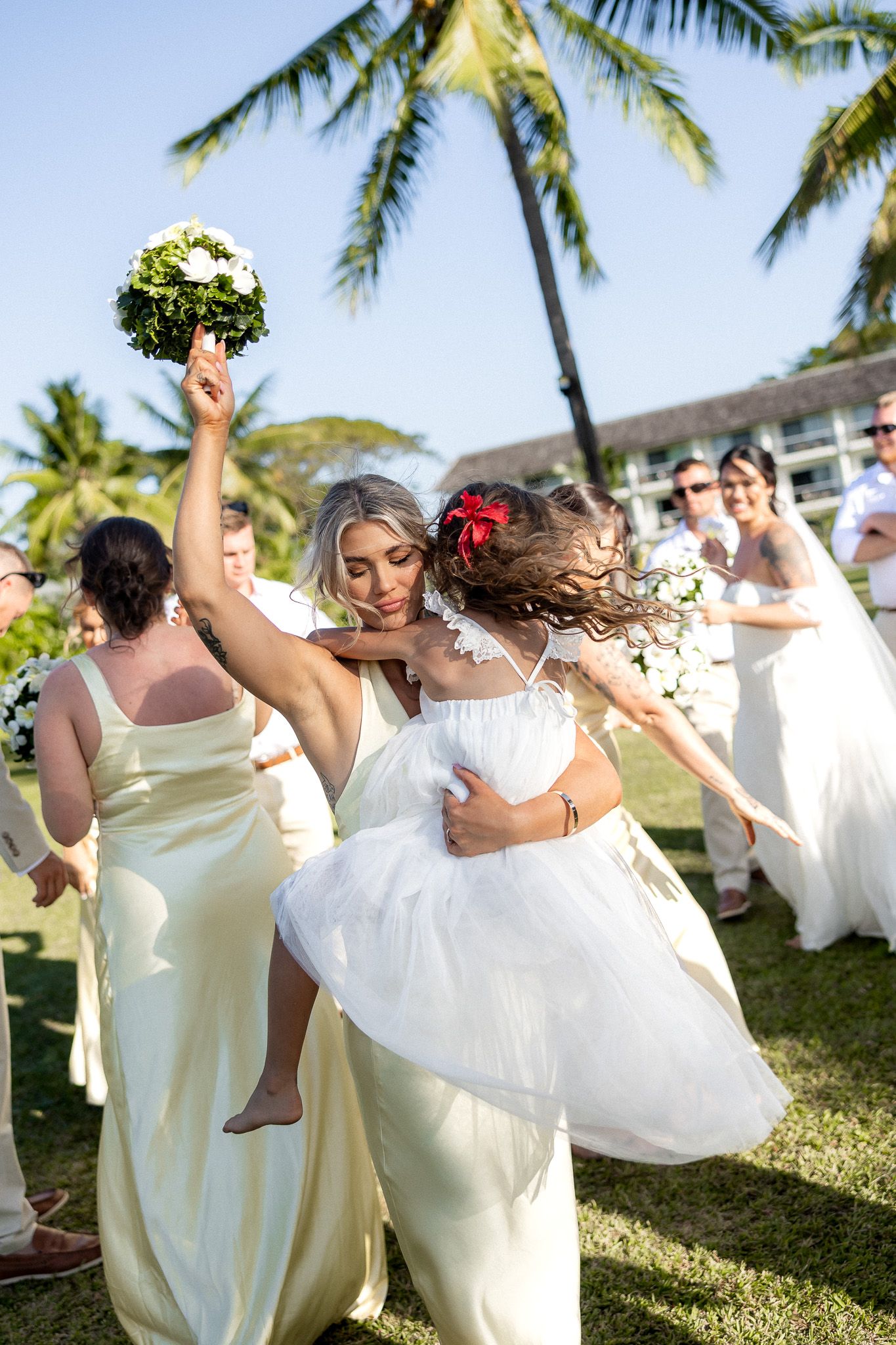 Bride holding bouquet high while hugging flower girl with bridesmaids in yellow dresses at outdoor wedding.