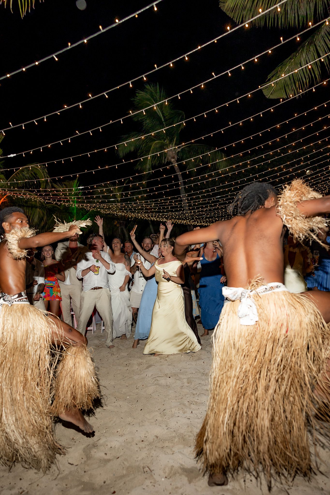 Fijian cultural dancers in grass skirts perform for wedding guests under string lights on beach.