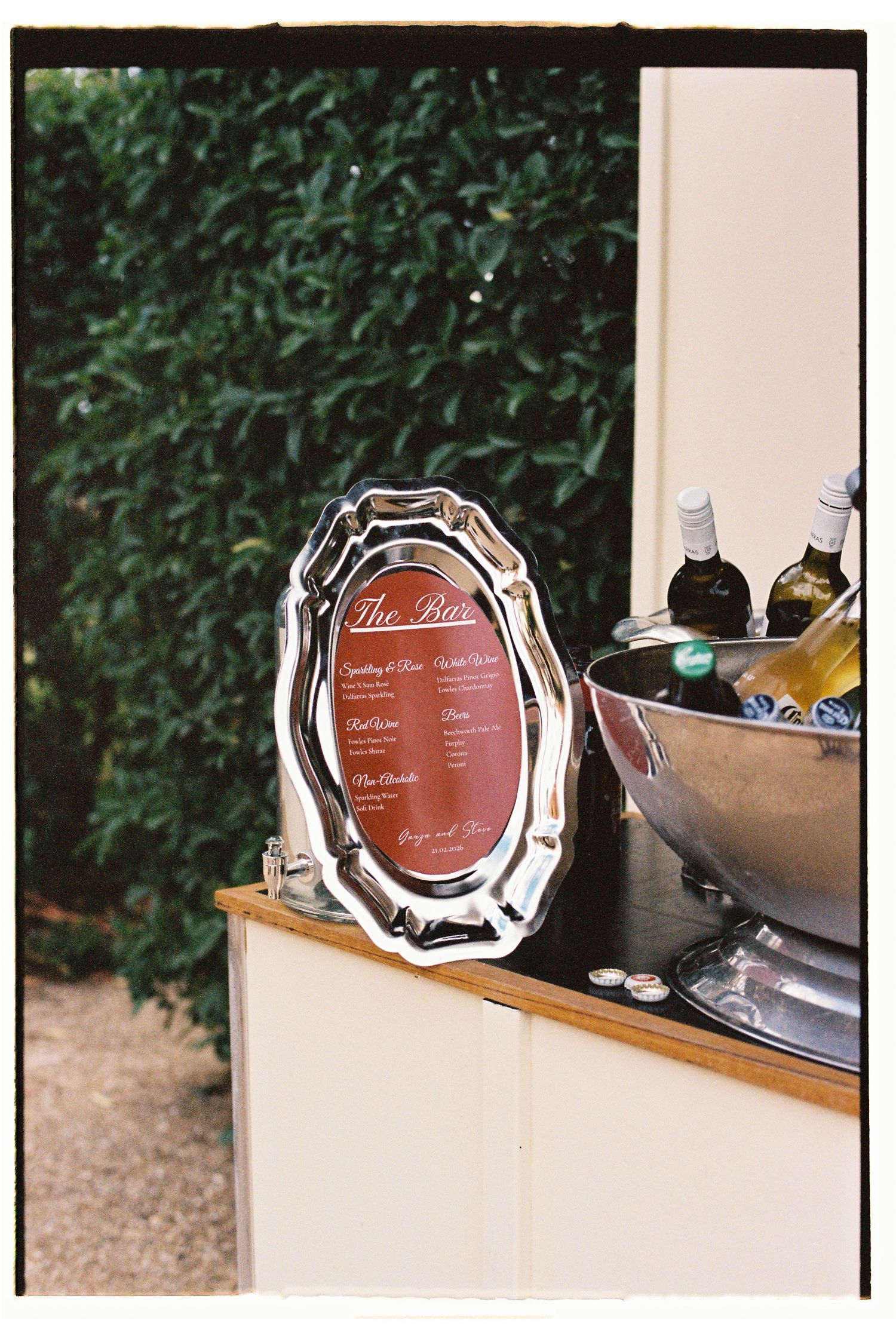 Elegant silver framed bar menu display beside wine bottles chilling in ice bucket at outdoor wedding reception.