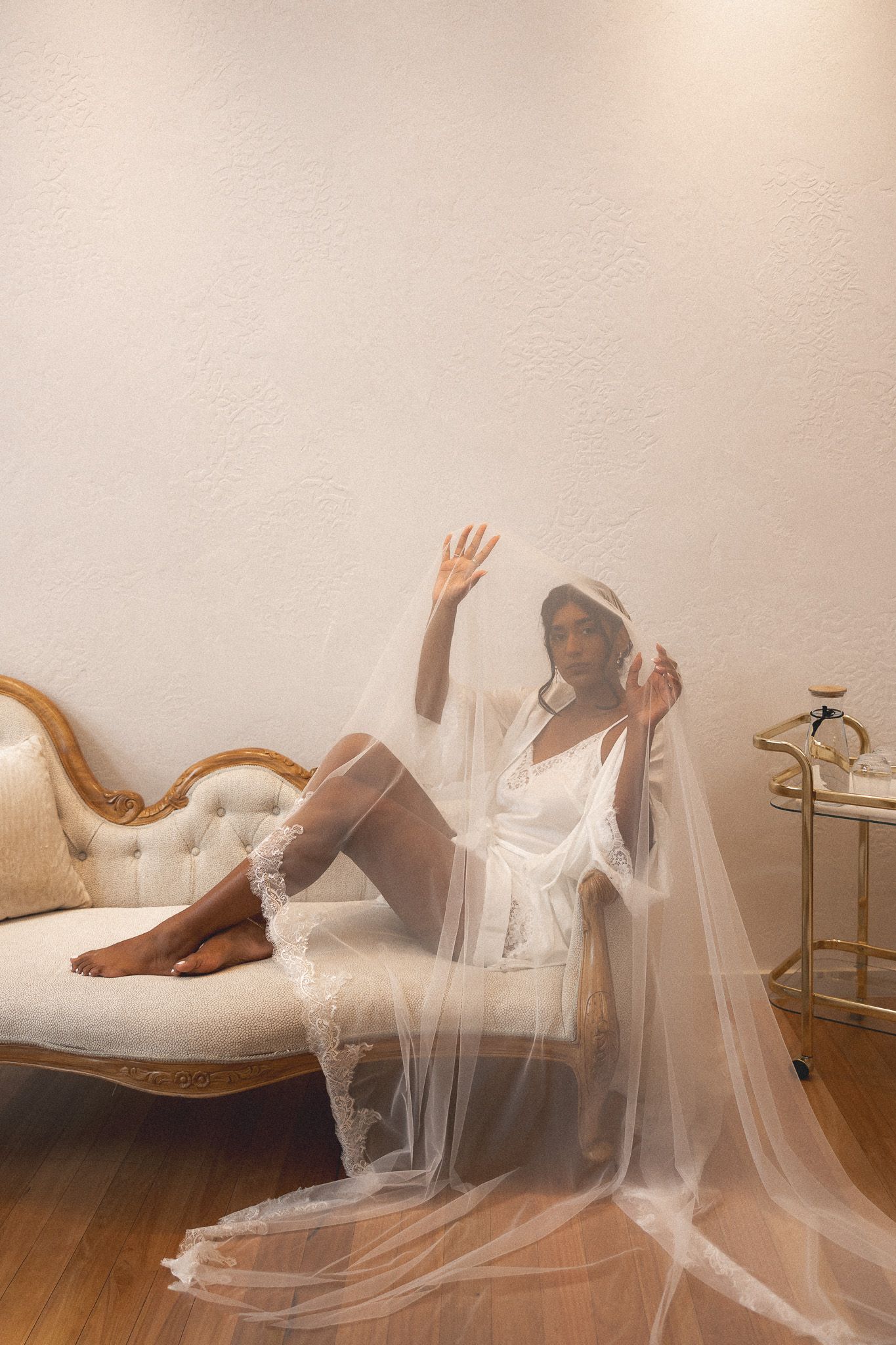 Bride in white robe and veil posing gracefully on vintage chaise lounge in soft lit room.
