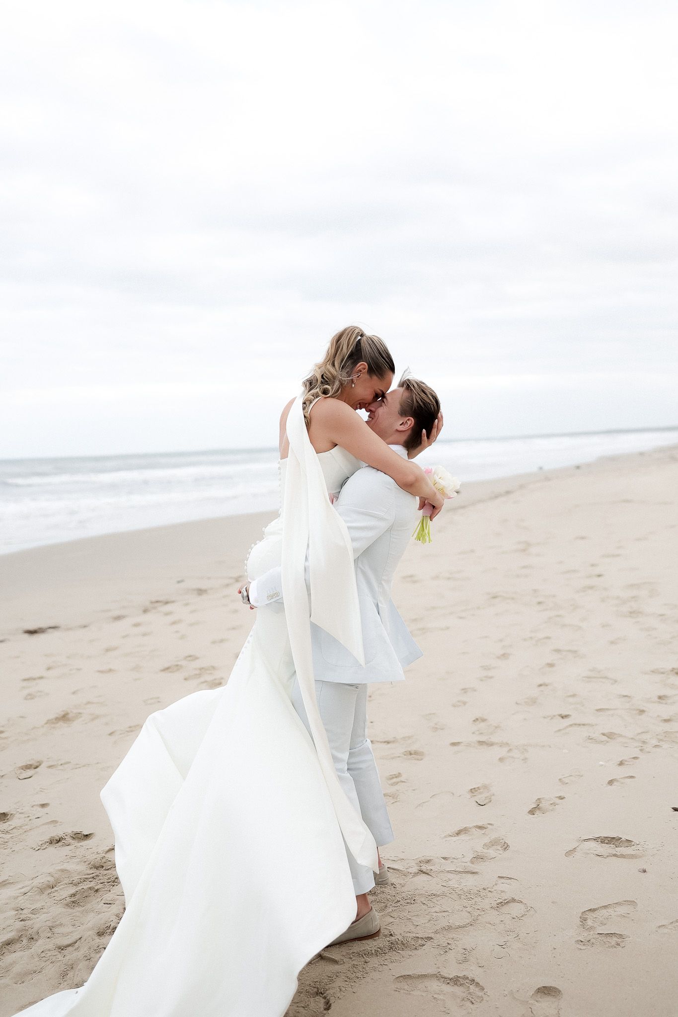 Groom lifting bride in white wedding dress on a sandy beach with ocean waves behind them.