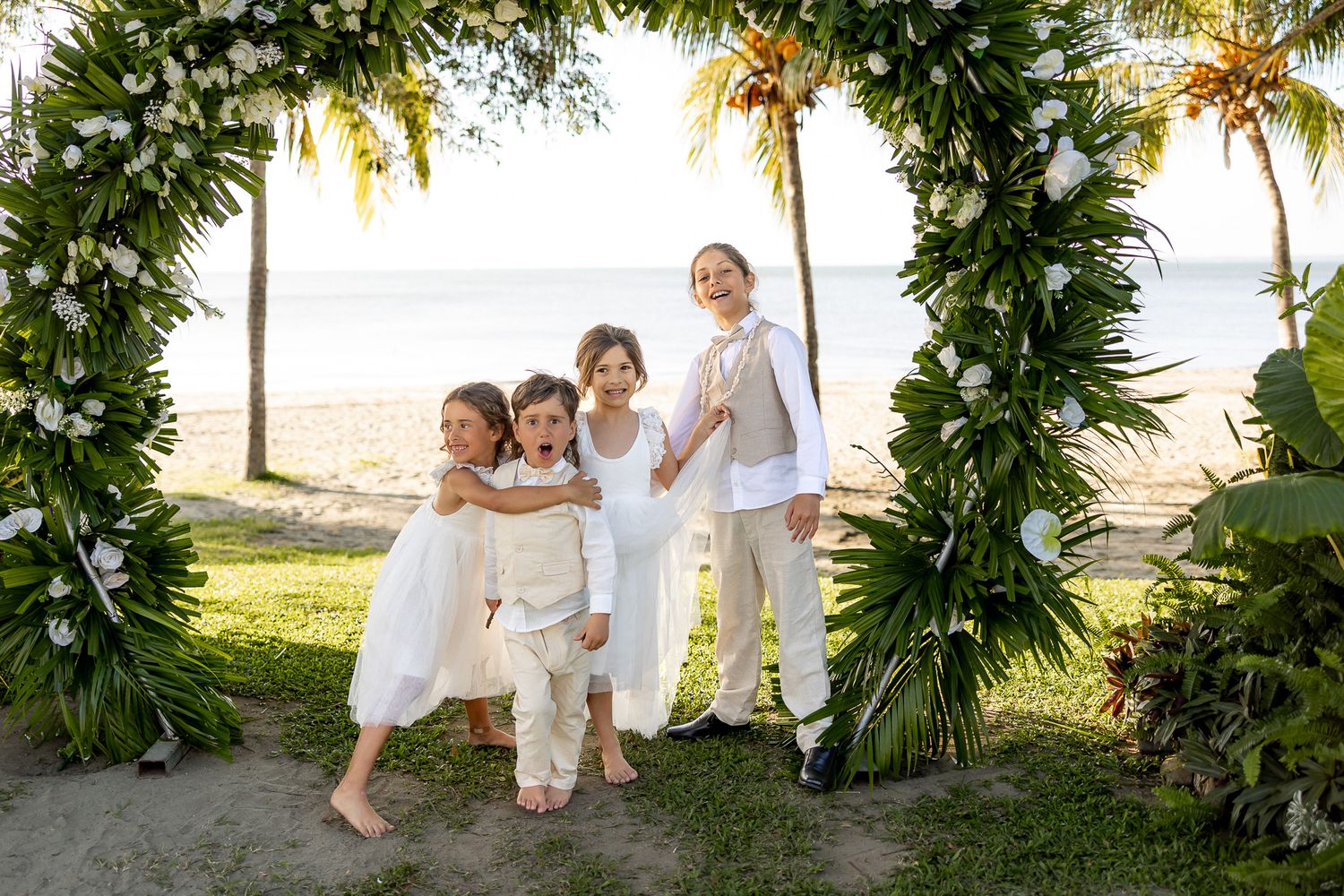 Four children in white outfits pose under a floral arch at a beach wedding ceremony.