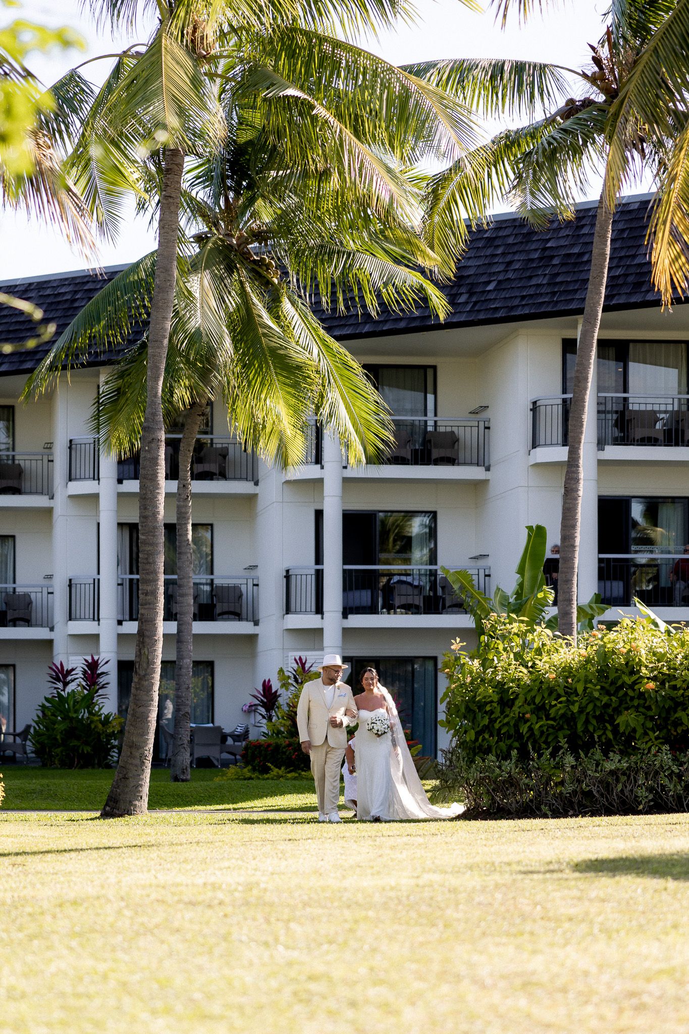 Bride and groom walking hand in hand on tropical resort lawn surrounded by palm trees and white building.