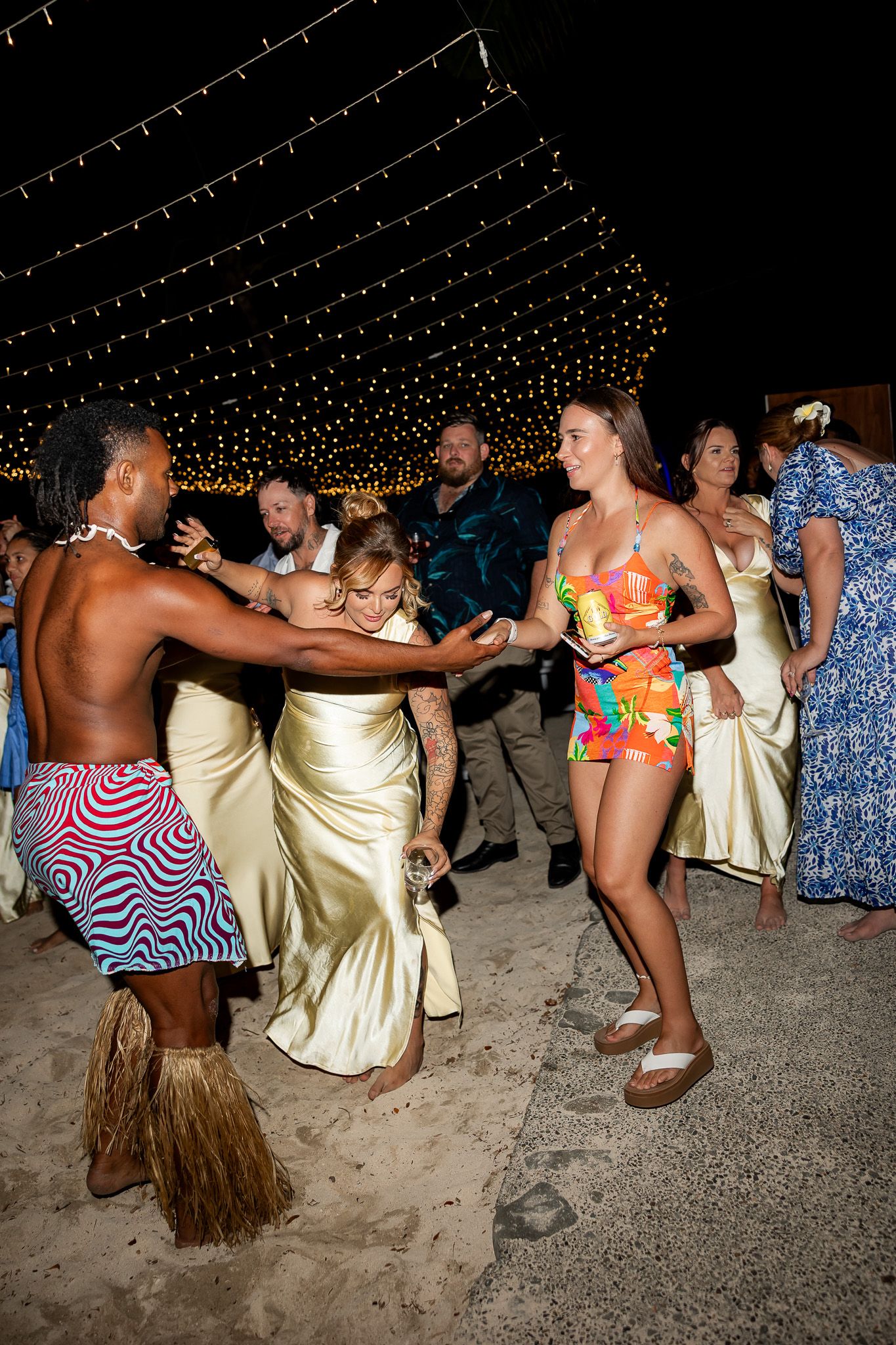 Fijian dancer in colorful sarong dances with wedding guests under string lights at reception.