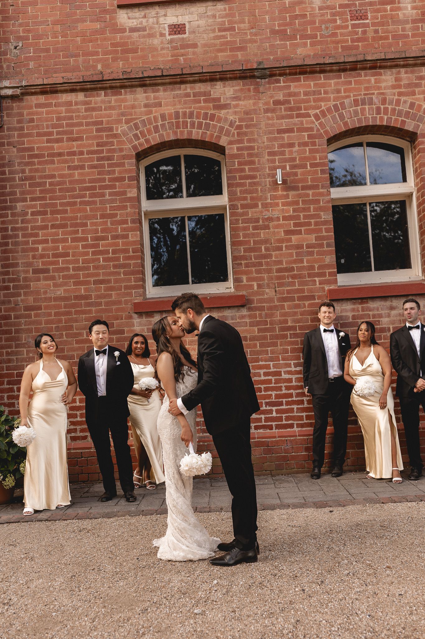 Bride and groom share a romantic kiss while wedding party stands before a red brick wall.