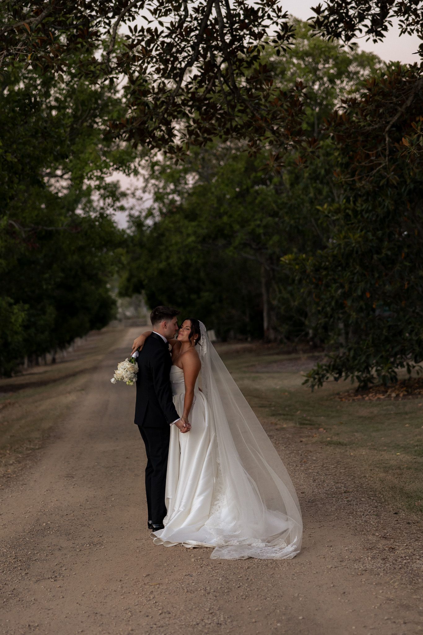 Bride and groom kissing on a tree-lined dirt path at sunset on their wedding day.