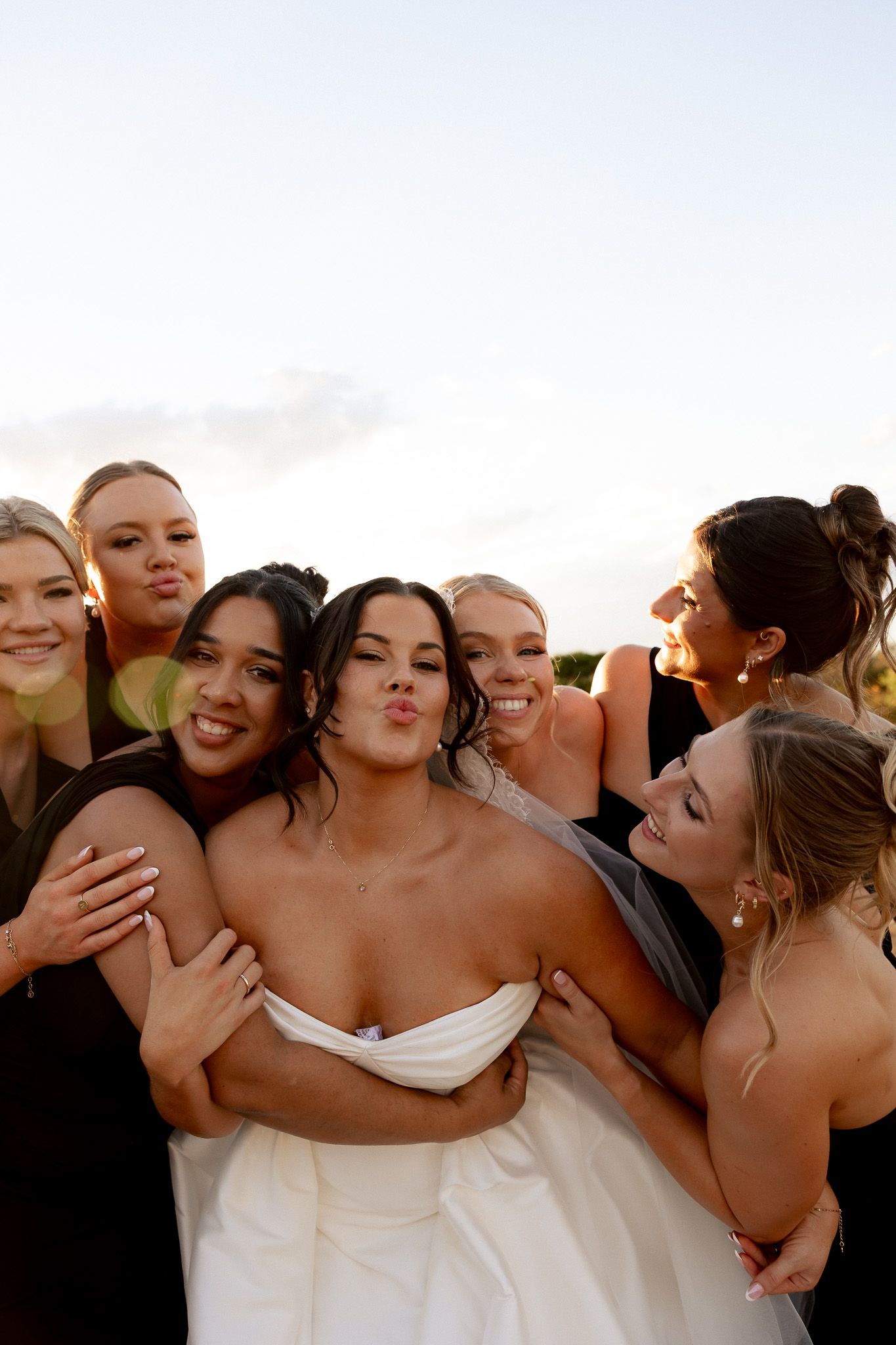 Bridesmaids in black dresses hugging and kissing bride in white strapless gown during fun outdoor wedding photo.