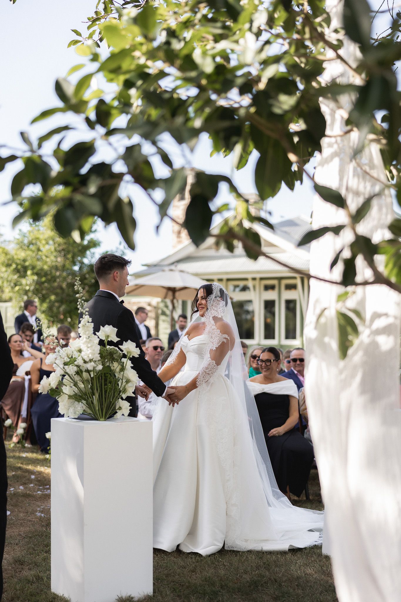 Bride and groom holding hands at outdoor wedding ceremony framed by lush green leaves.