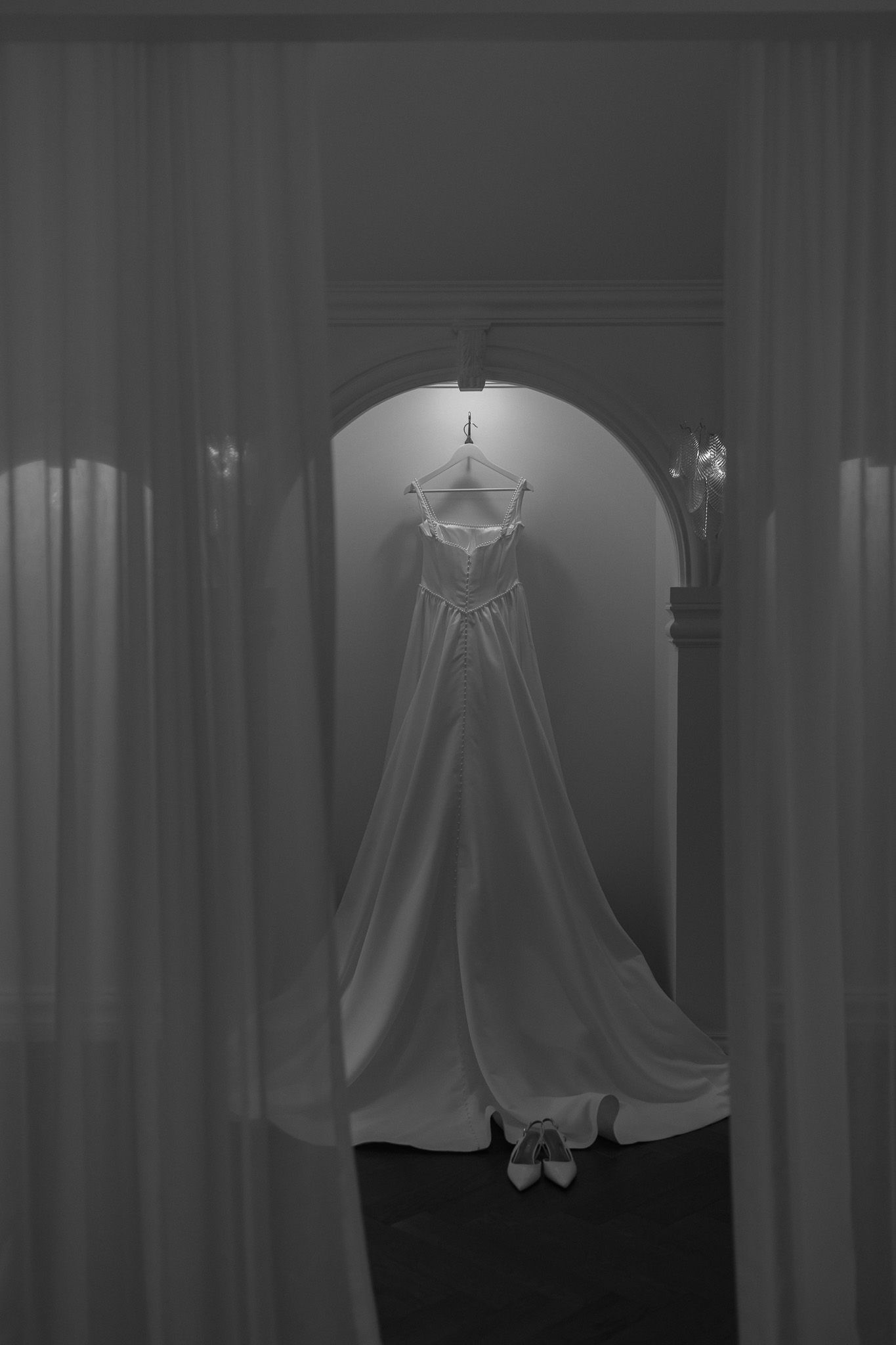 Black and white photo of an elegant wedding dress hanging in an arched alcove, with sheer curtains framing the scene.
