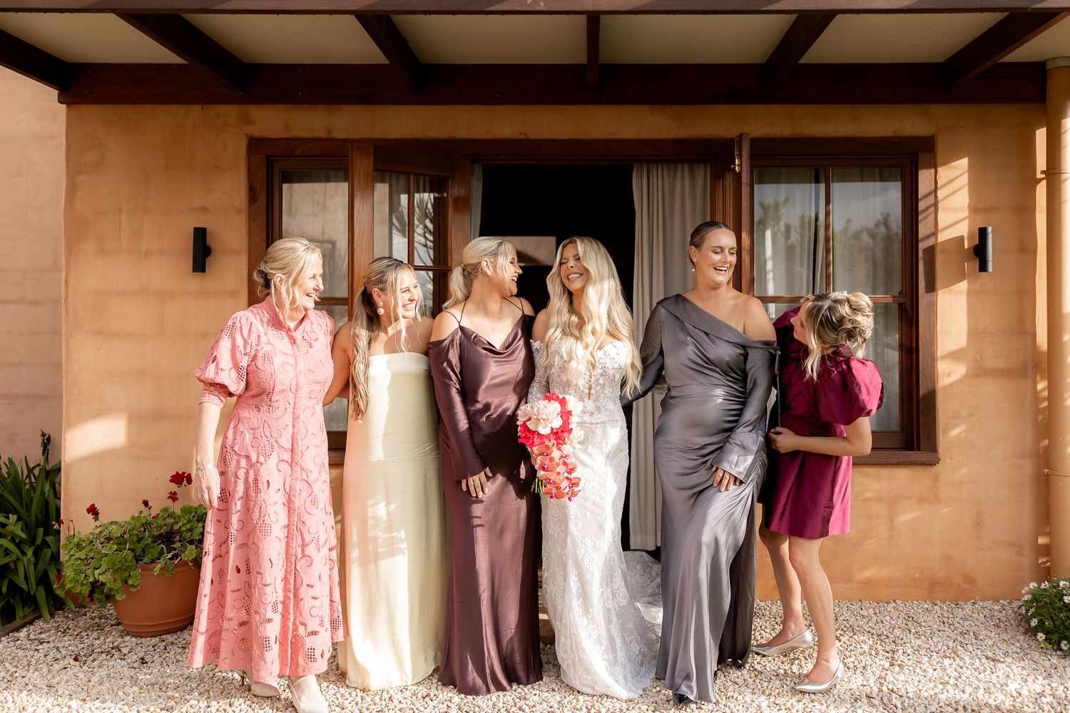 Bride with bridesmaids and family in elegant dresses posing outside a rustic venue on a sunny wedding day.