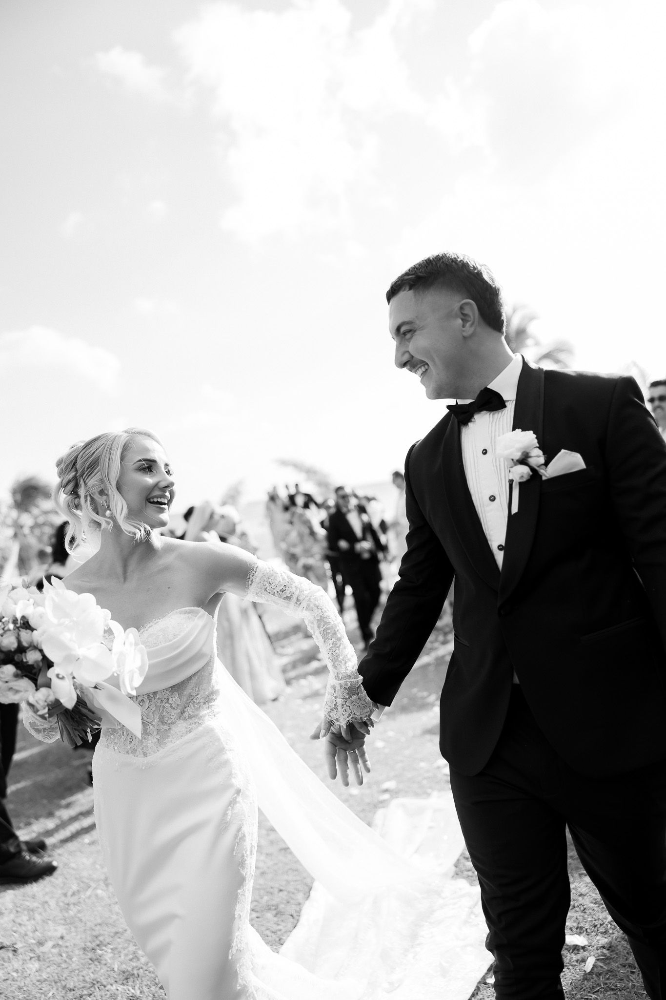 Black and white photo of a joyful bride and groom holding hands and smiling at each other on their wedding day.