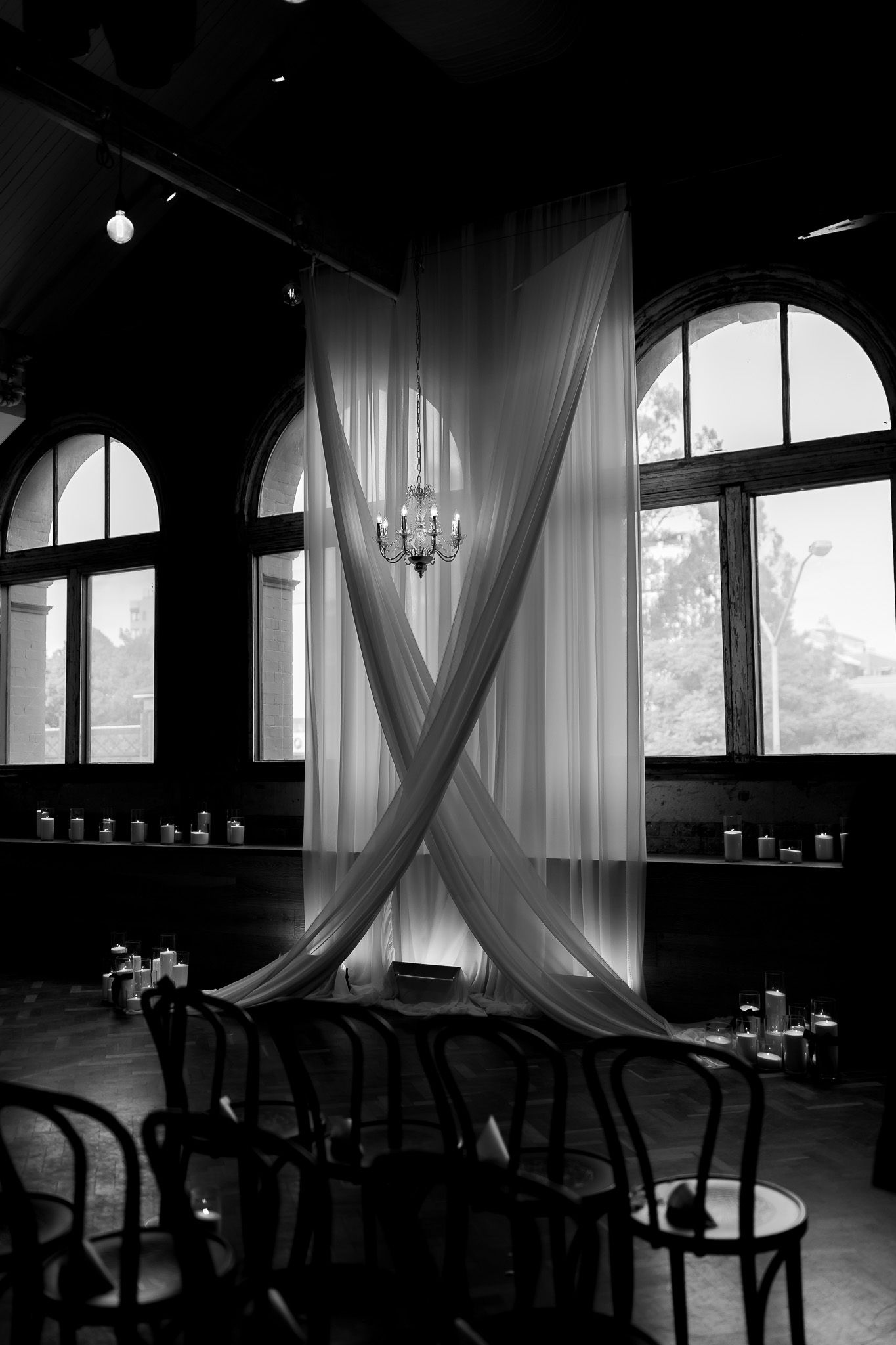 Black and white photo of elegant wedding ceremony setup with draped fabric backdrop, chandelier, and Thonet chairs in historic venue.