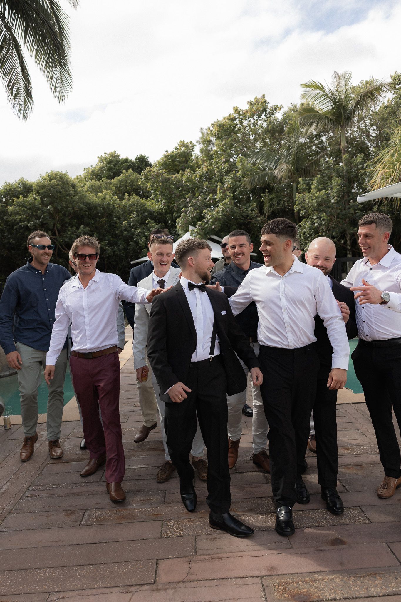 Groom in tuxedo celebrates with groomsmen in white shirts near outdoor pool surrounded by palms.