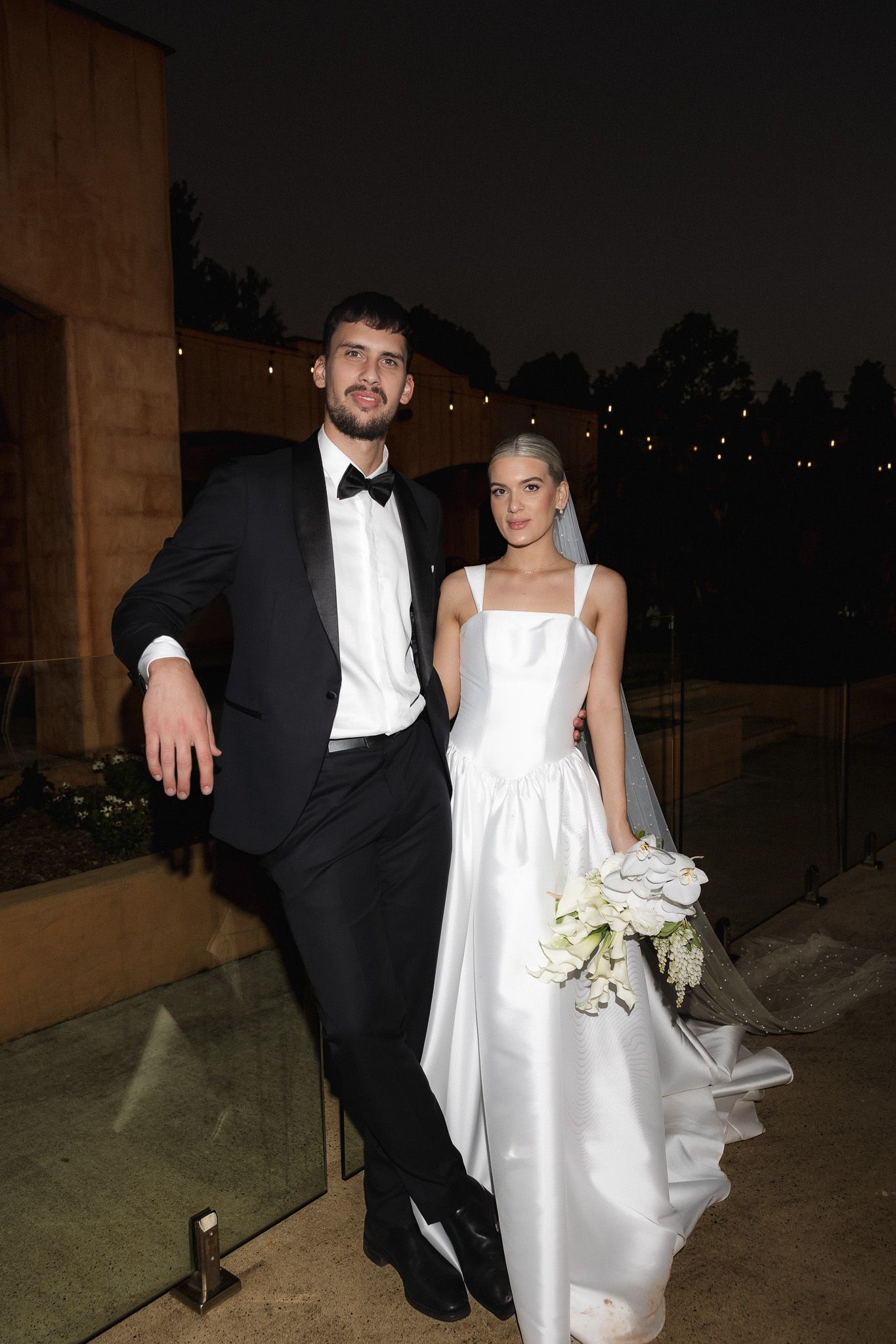 Elegant bride and groom pose at night, groom in black tuxedo and bride in white satin gown with bouquet.