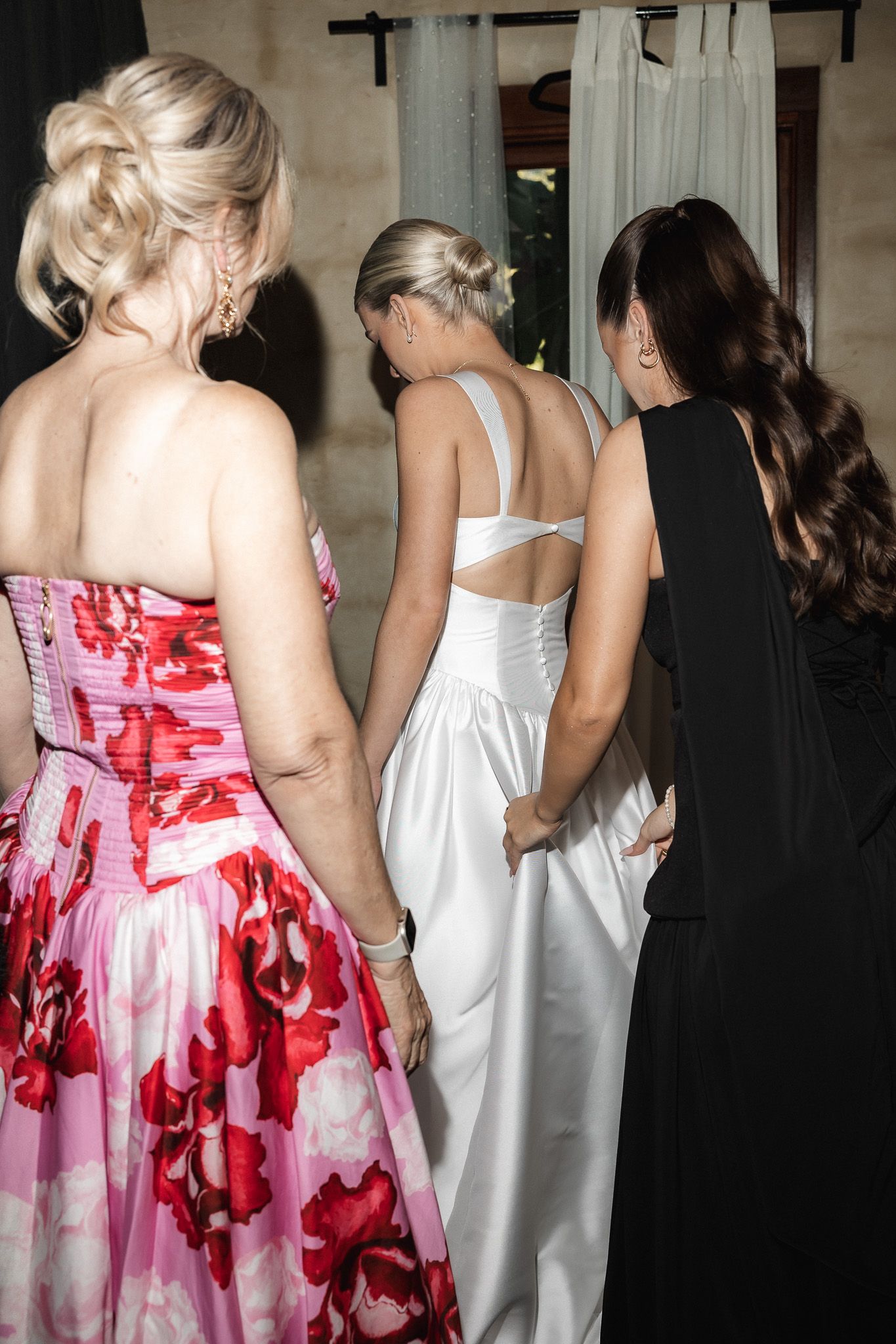 Three women in formal gowns, including a bride in white, stand together during wedding preparations.