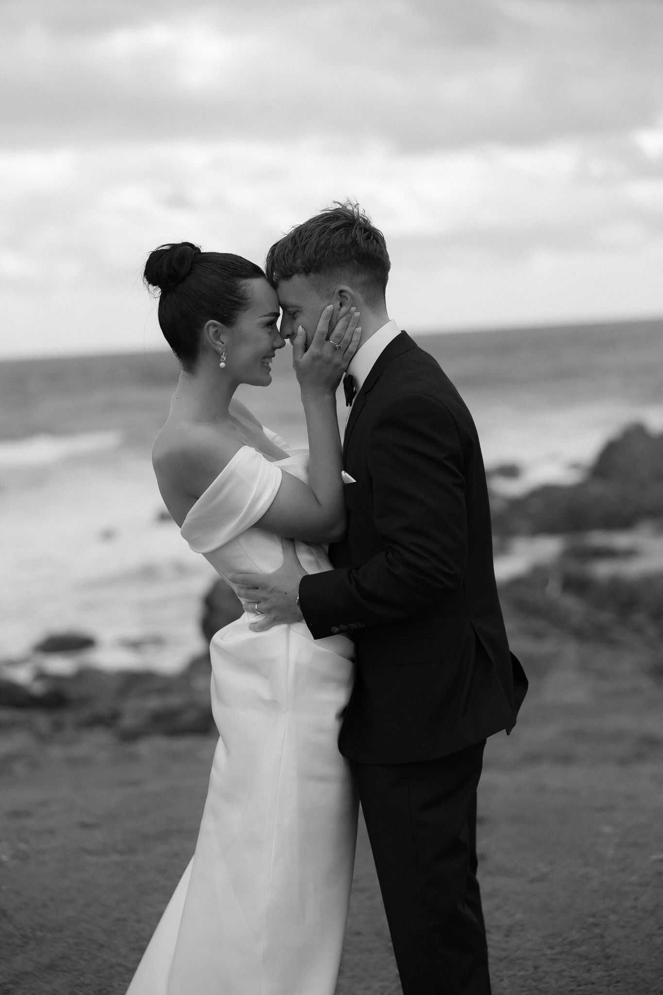 Black and white photo of a bride and groom embracing near the ocean, sharing an intimate moment in wedding attire.