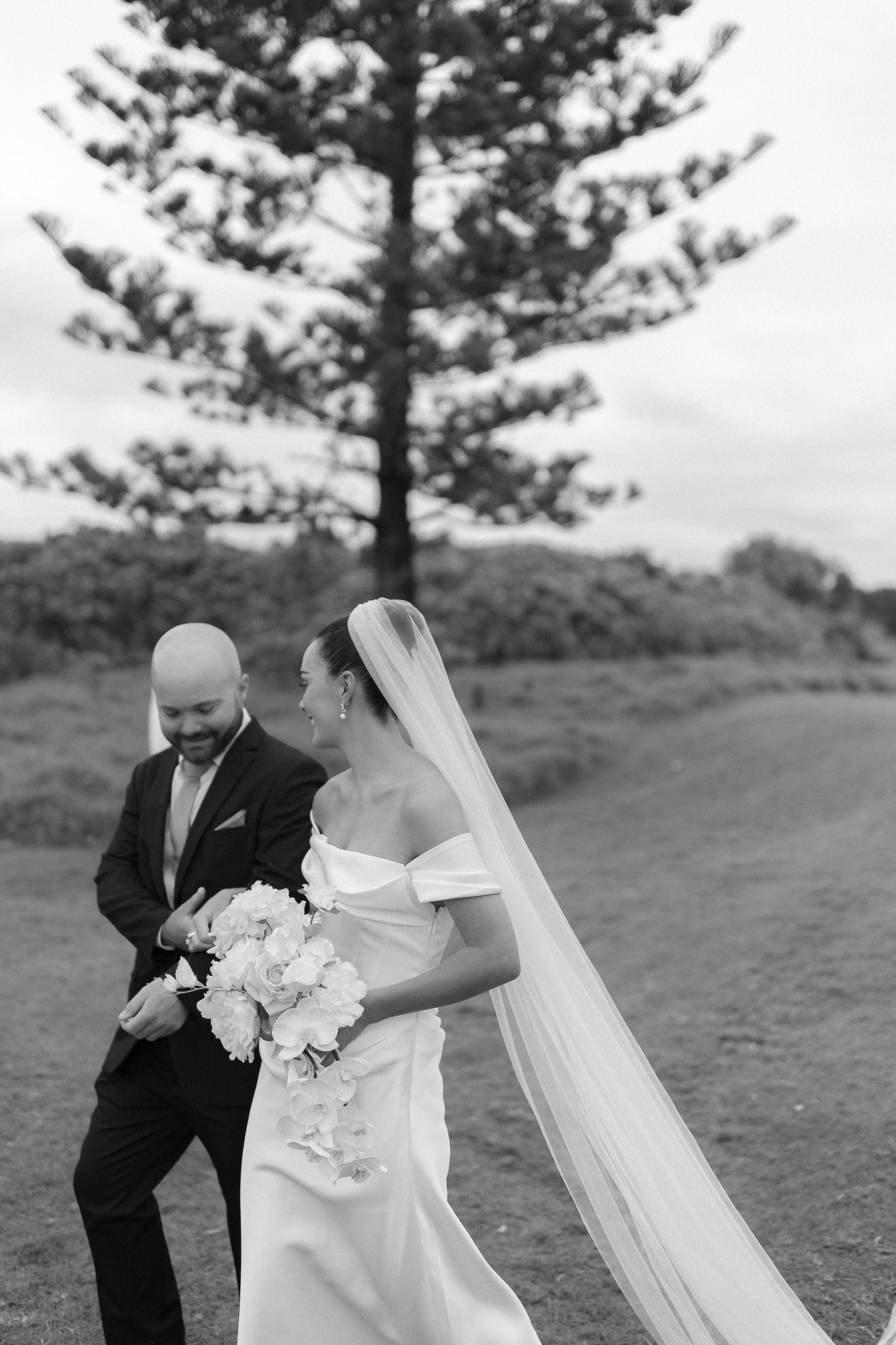 Black and white photo of a smiling bride in off-shoulder gown with veil and groom in suit walking outdoors.
