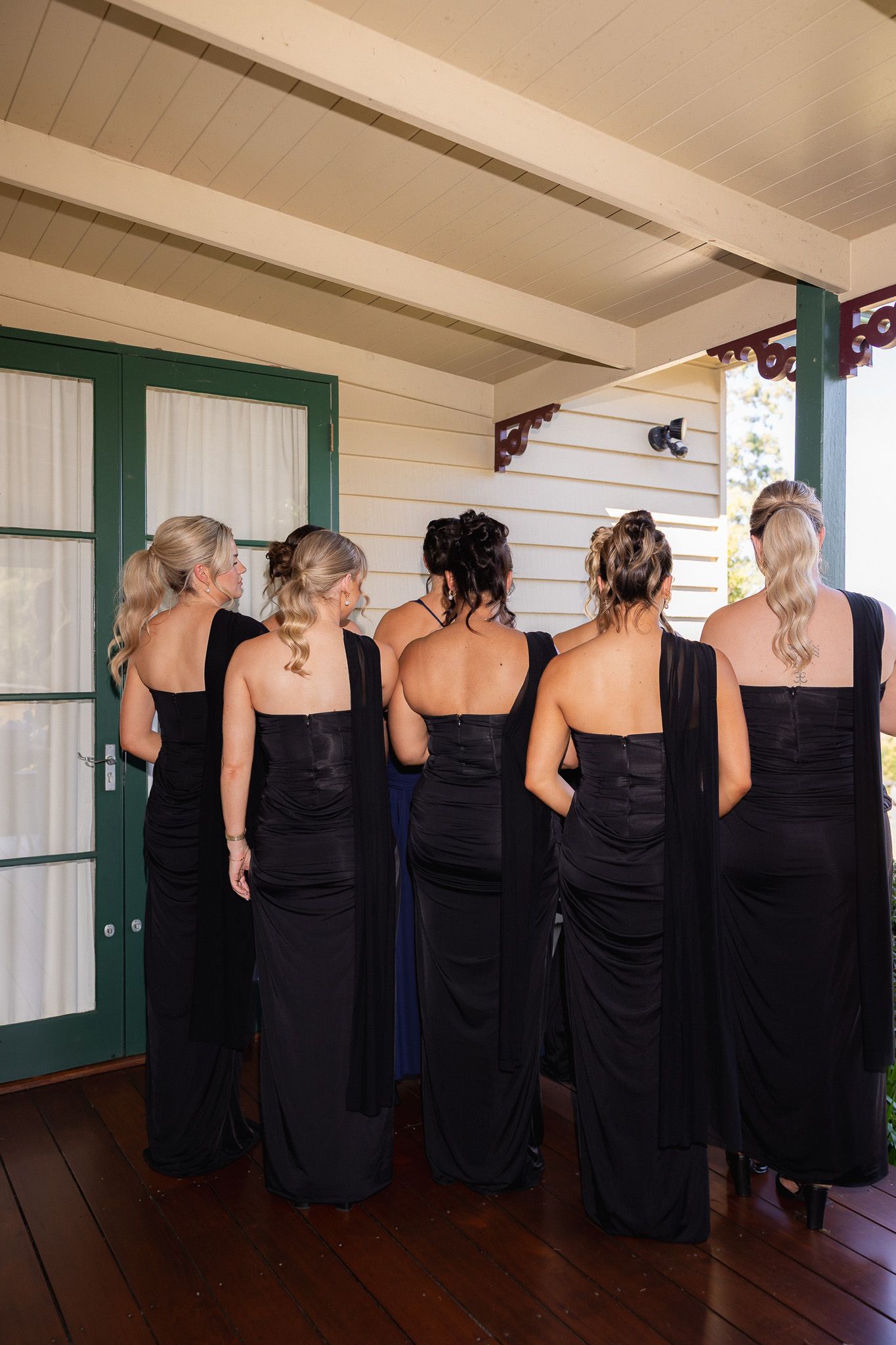 Five bridesmaids in elegant black gowns standing with backs to camera on a wooden porch.