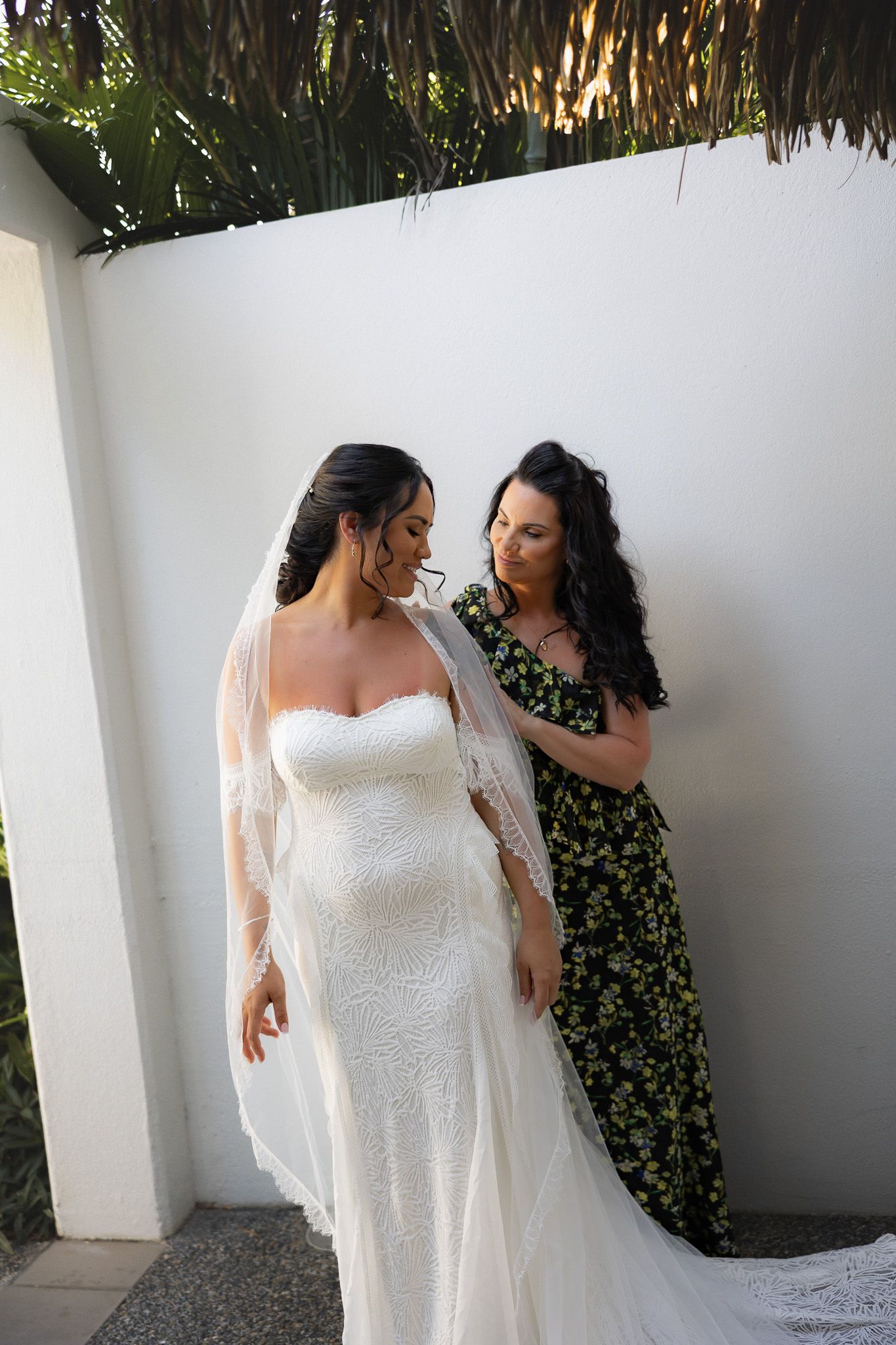 Helper adjusts bride's lace wedding dress and veil against white wall in tropical setting.