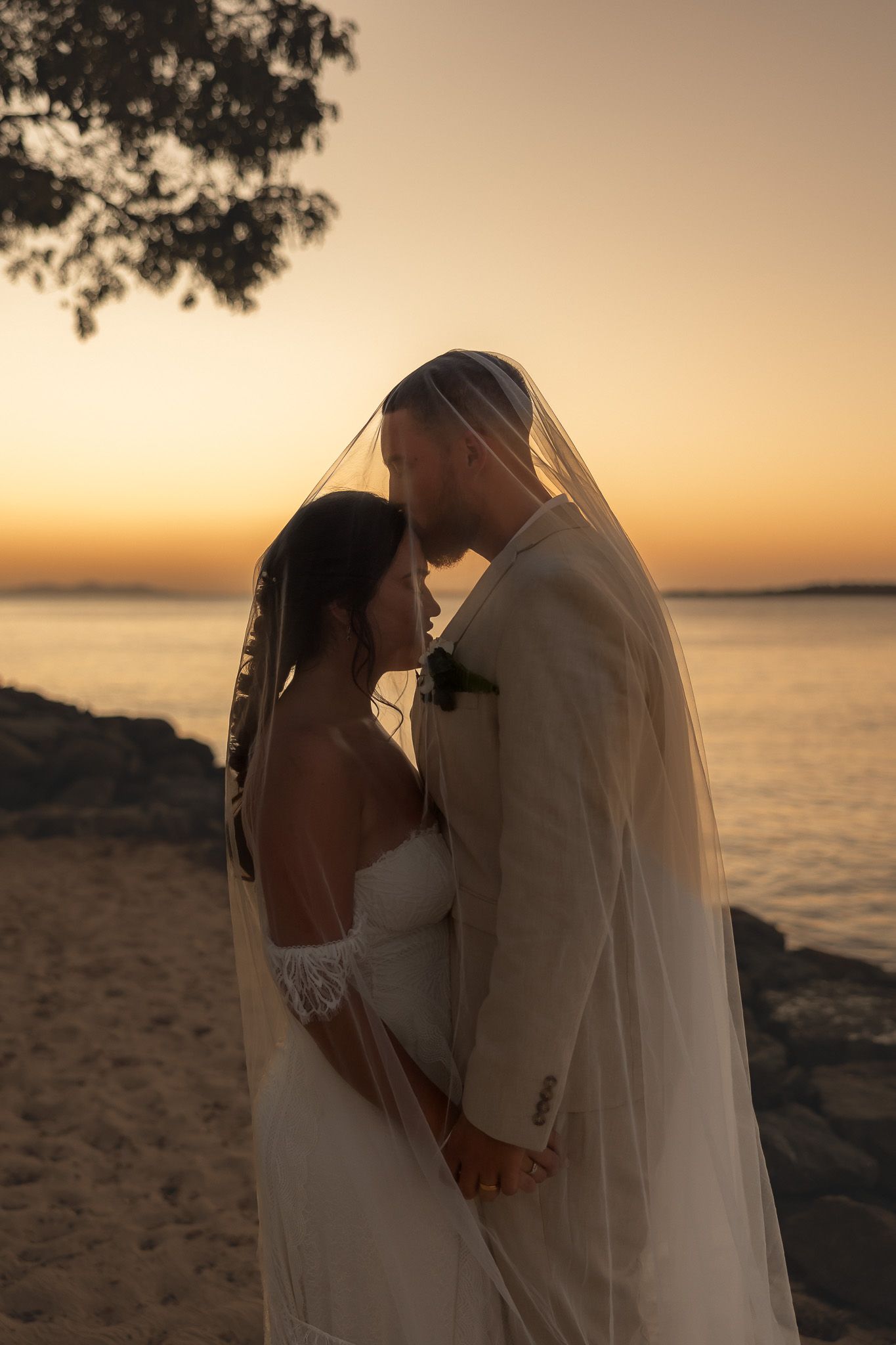 Bride and groom share a romantic kiss under a veil at sunset on a tropical beach with golden sky.
