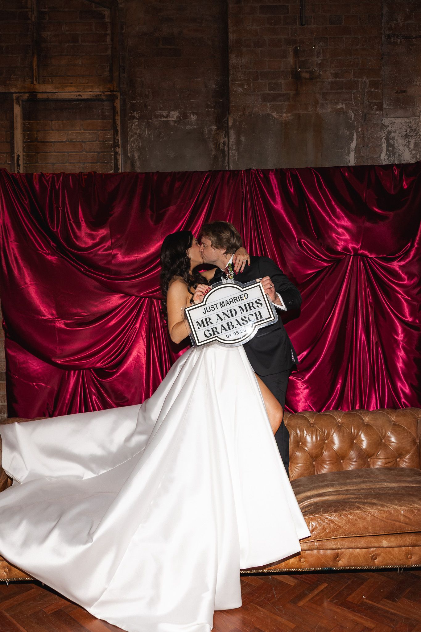 Newlyweds kissing and holding a Mr. and Mrs. sign in front of a red velvet curtain backdrop.