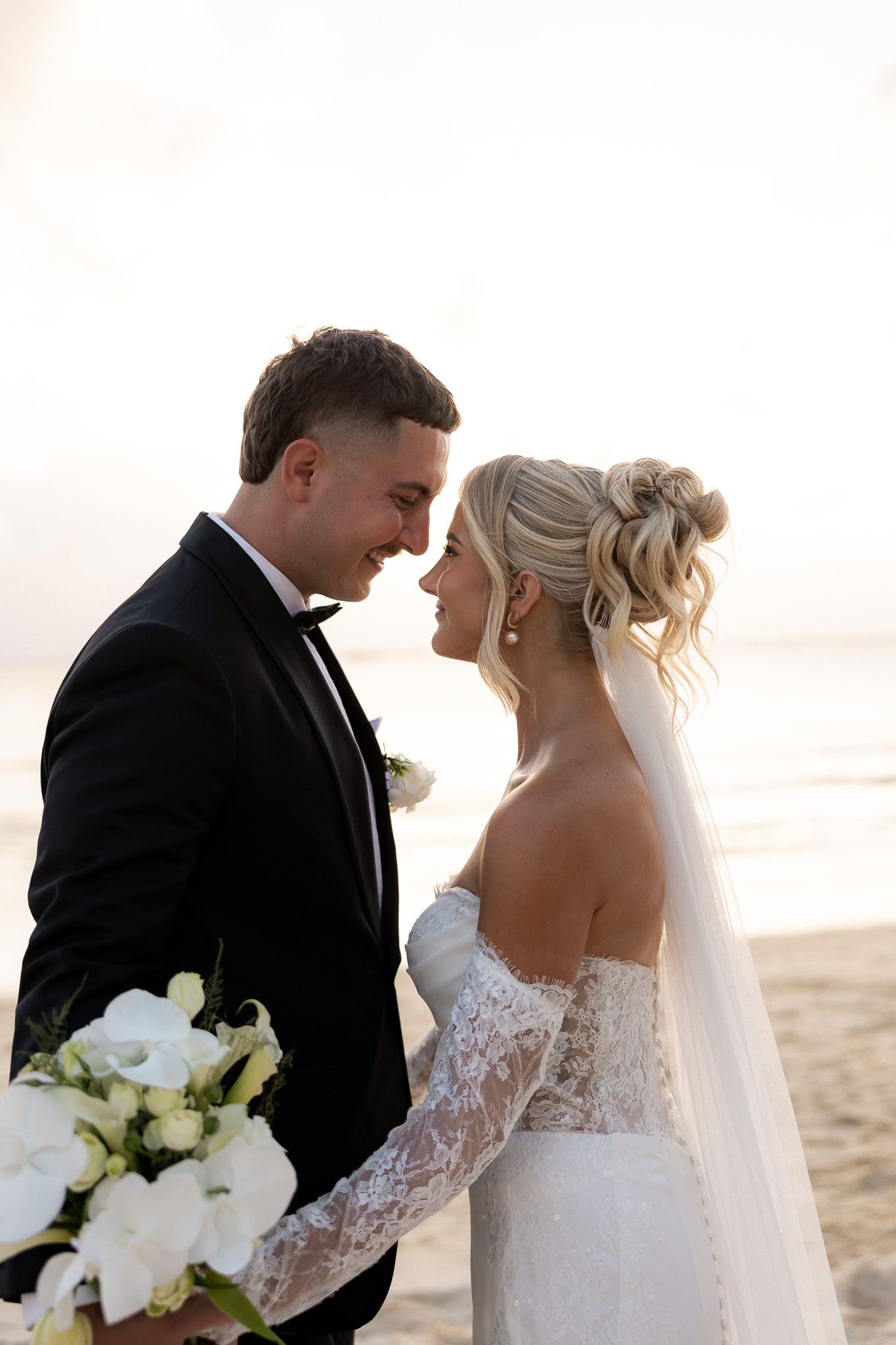 Bride and groom share a romantic moment on a beach at sunset, with bride in lace gown holding white floral bouquet.