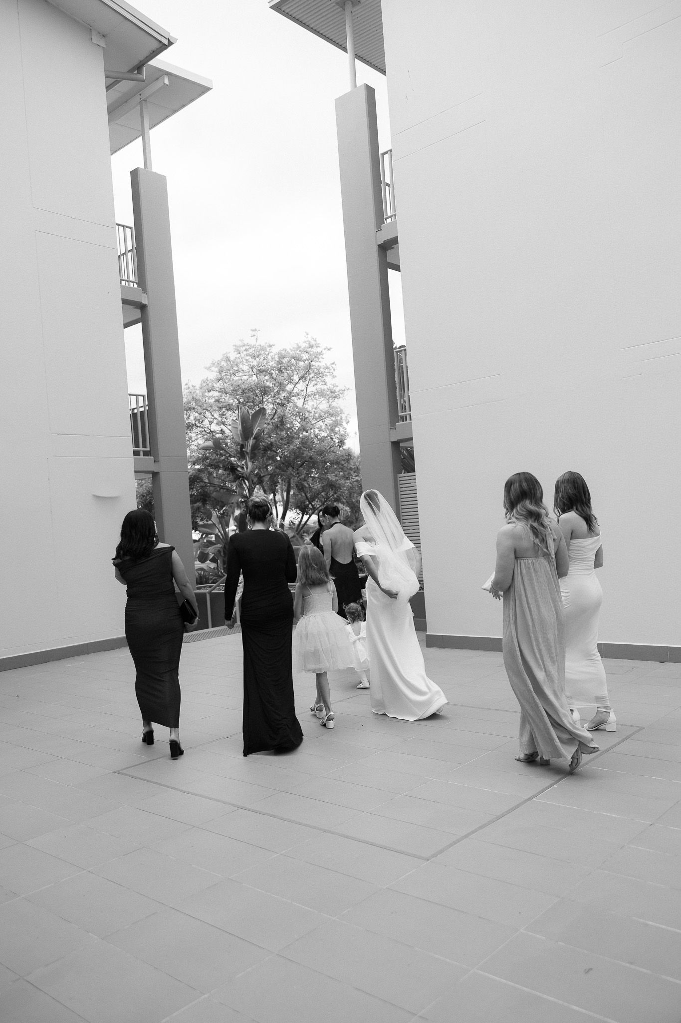 Black and white photo of bride in veil walking with bridesmaids through modern venue columns.