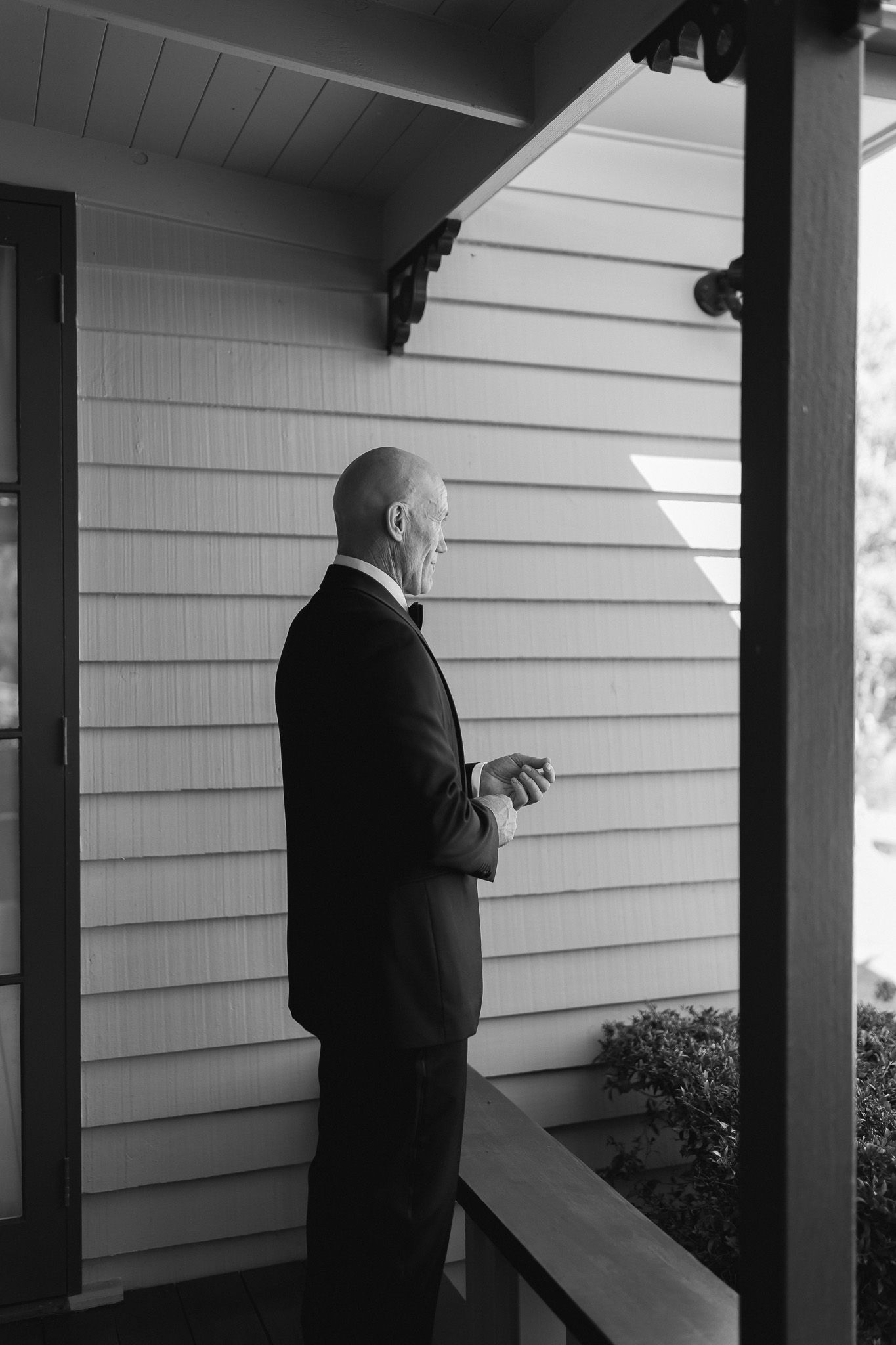 Bald man in tuxedo standing on wooden porch in black and white wedding photography.