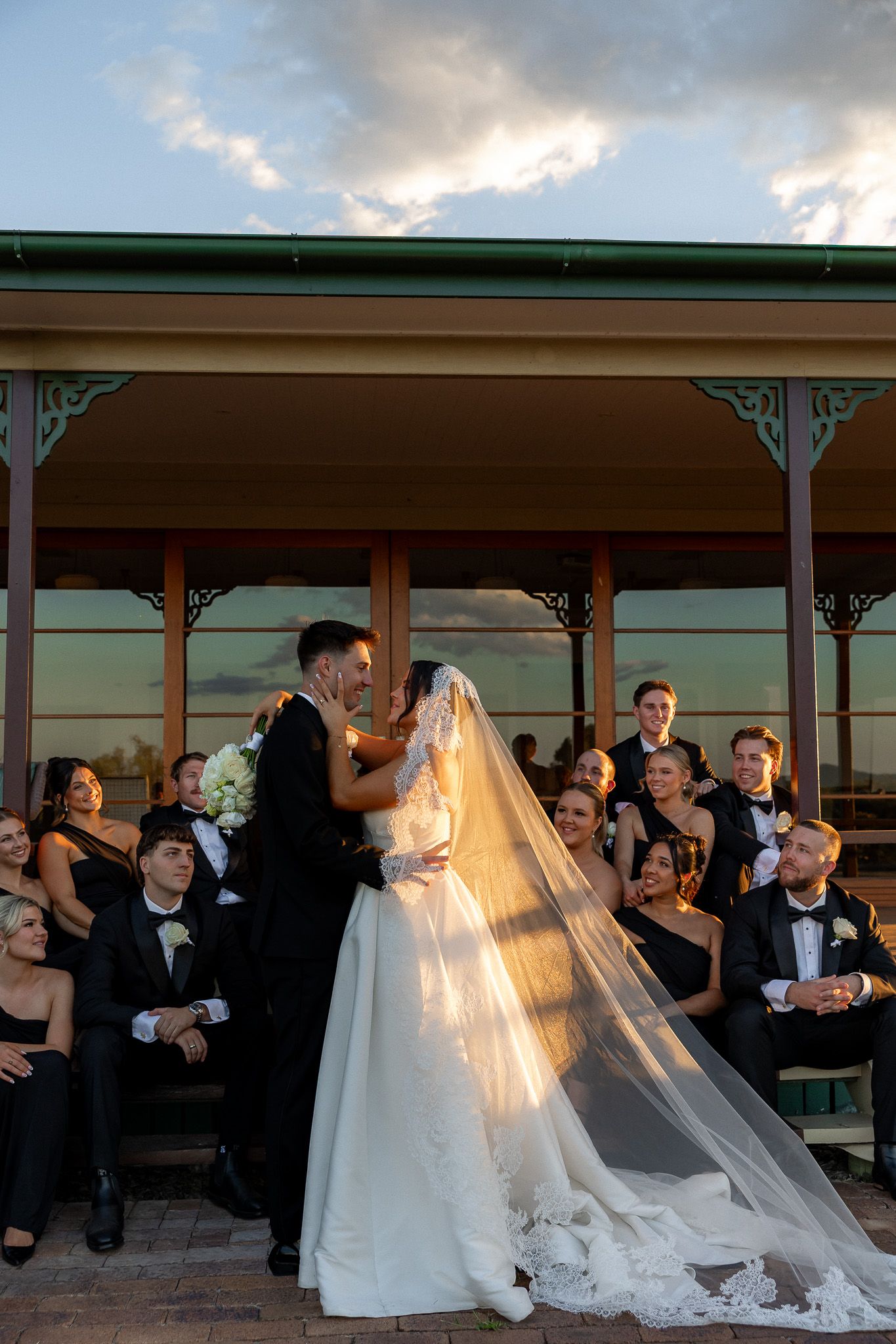 Bride and groom share a romantic moment with wedding party seated behind them at sunset venue.