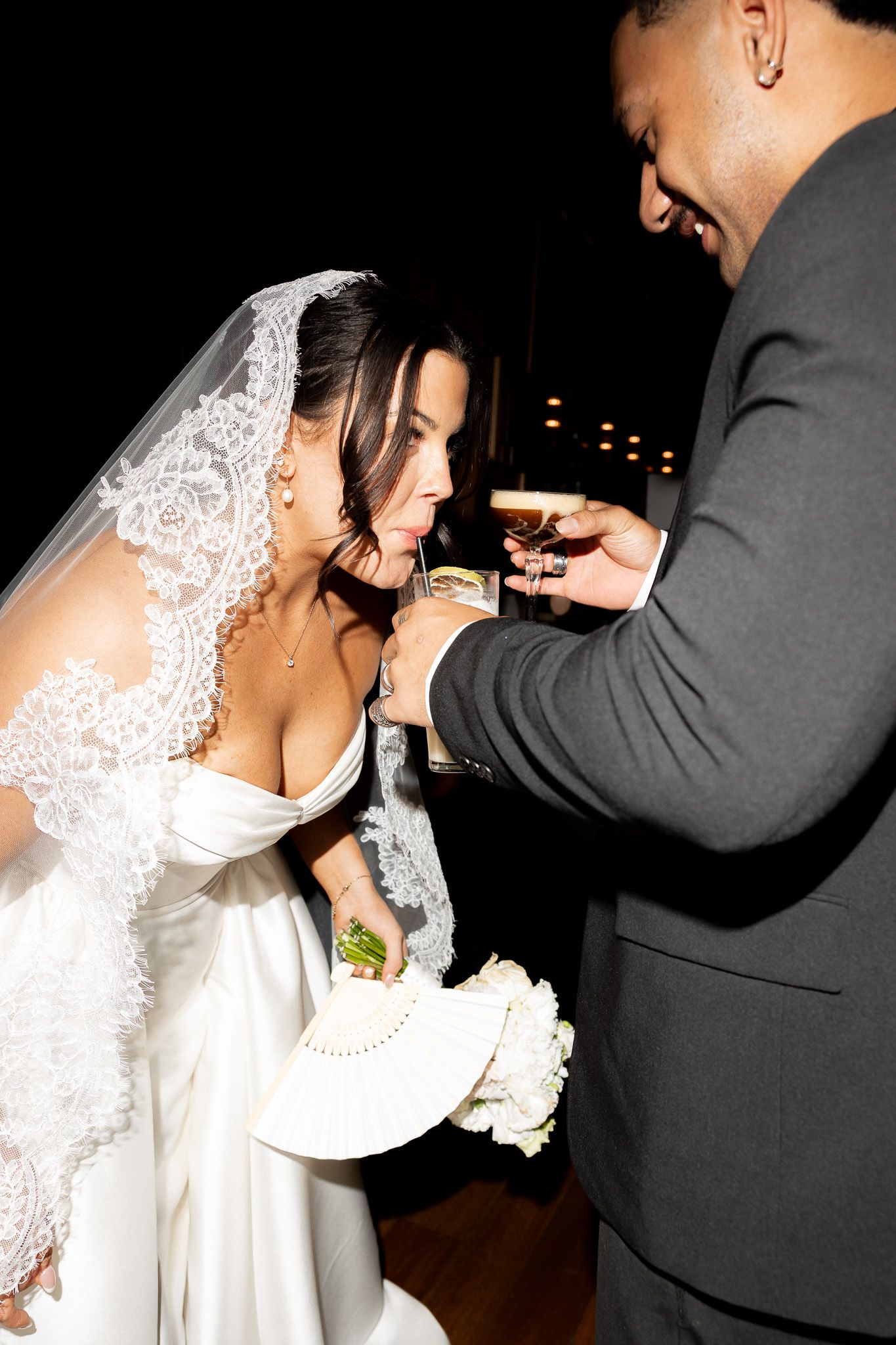 Bride in lace veil shares a drink with groom during evening wedding reception.
