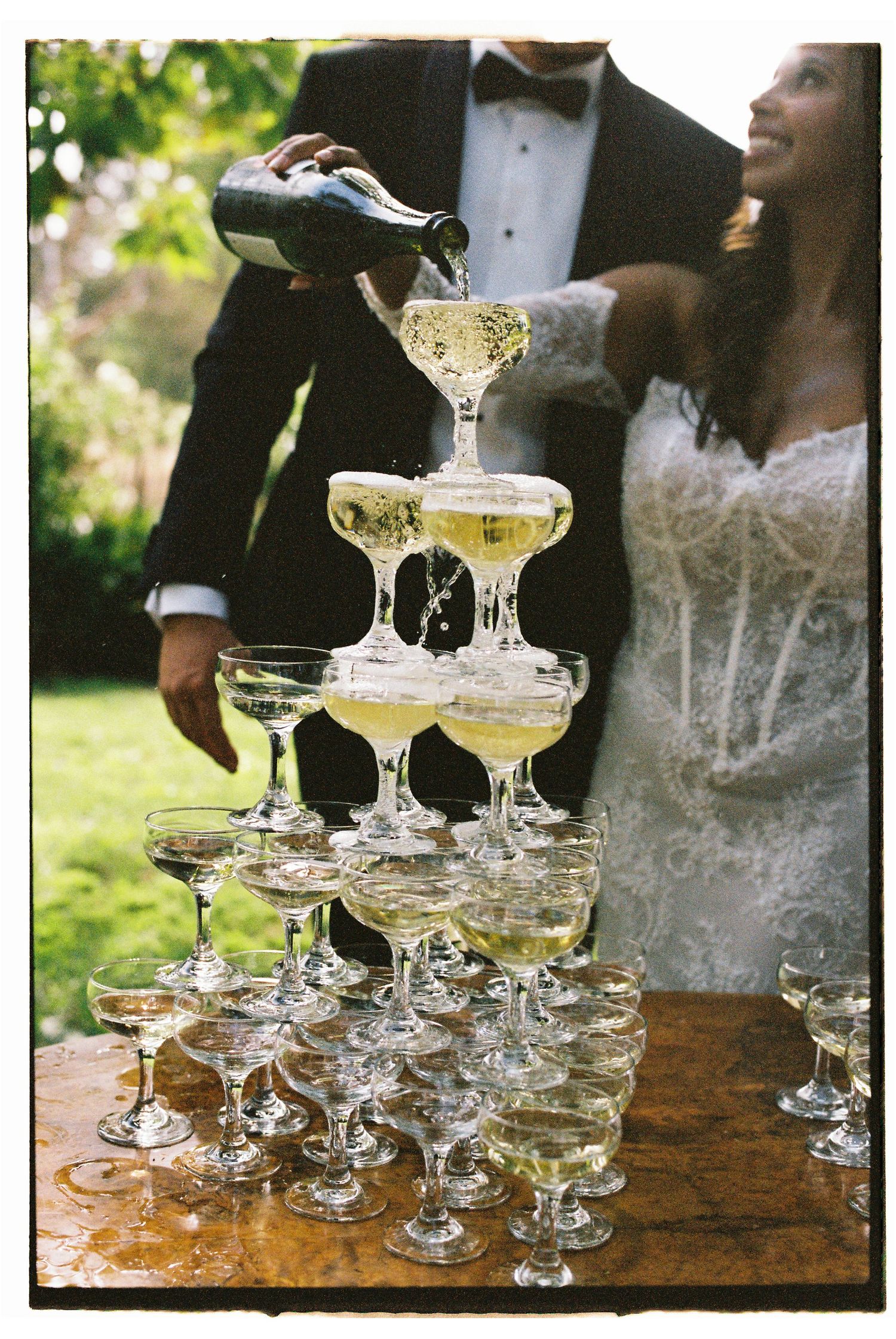 Bride and groom pouring champagne tower at outdoor wedding reception.