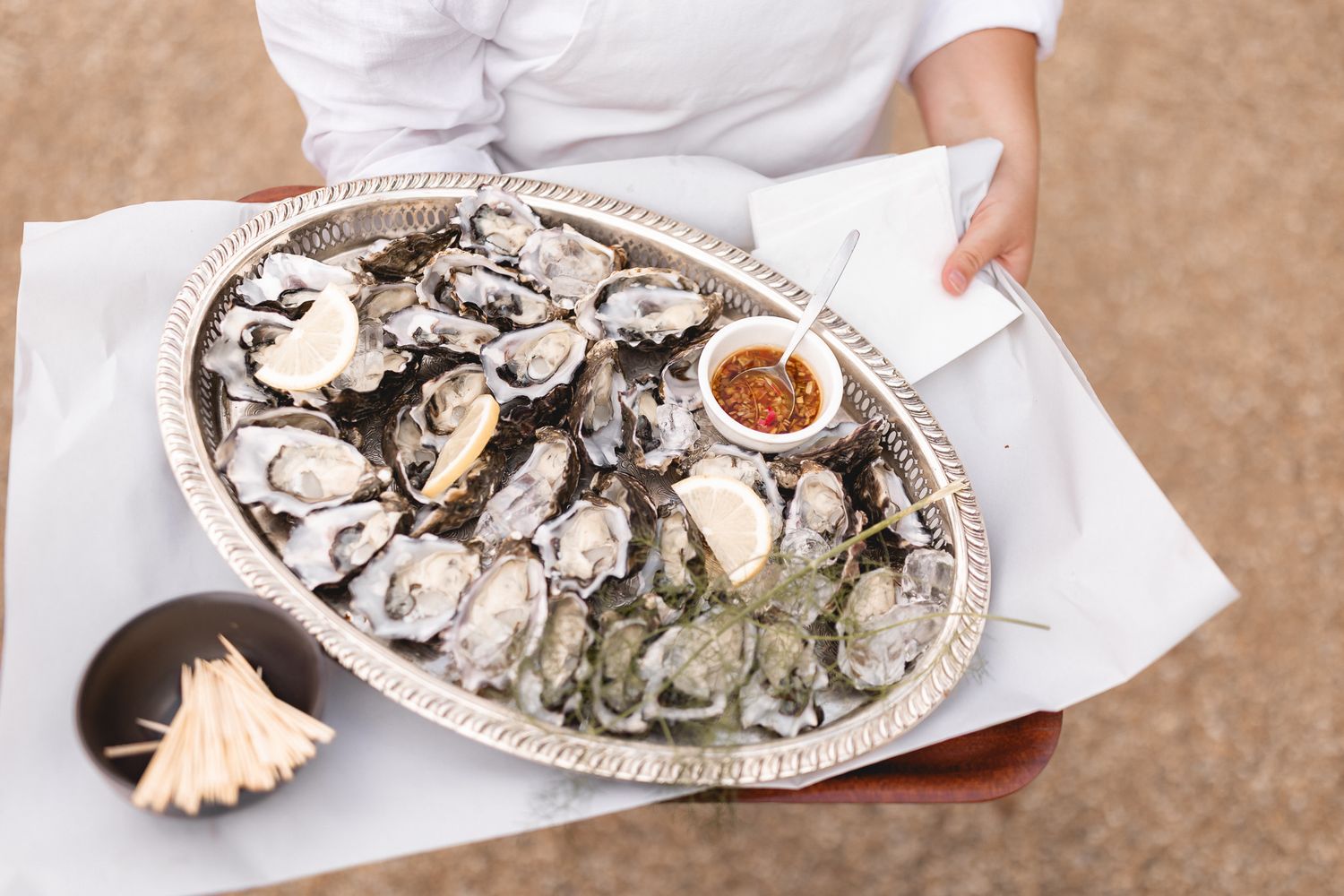Server holding a silver platter of fresh oysters on ice with dipping sauce at an outdoor event.