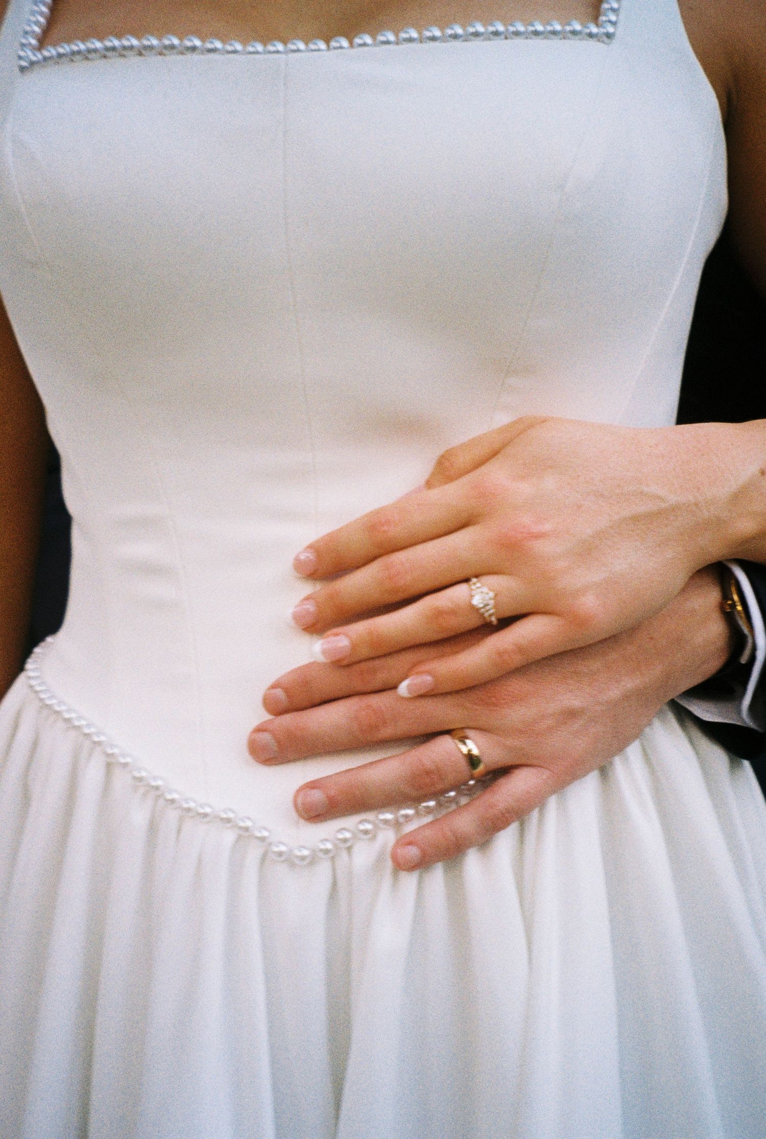Close-up of bride and groom's hands with wedding rings resting on white pearl-trimmed wedding dress.