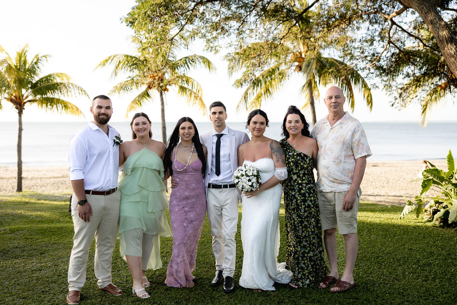 Wedding party group portrait on tropical beach lawn at golden hour with palm trees and ocean backdrop.