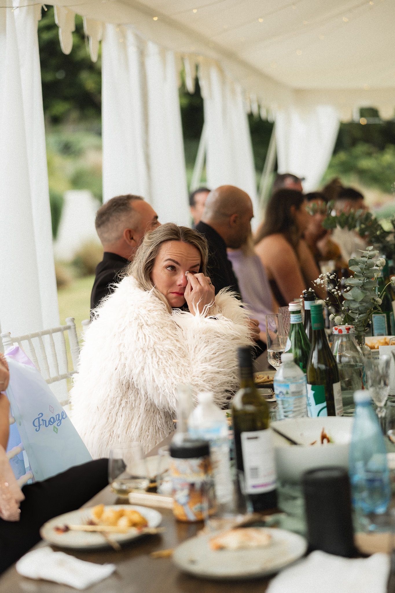 An emotional wedding guest in a white fur coat wiping tears at a reception dinner table.