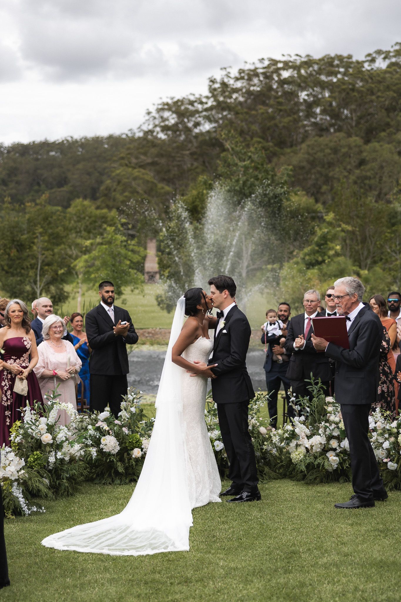 Bride and groom sharing first kiss at outdoor wedding ceremony with fountain and guests applauding.